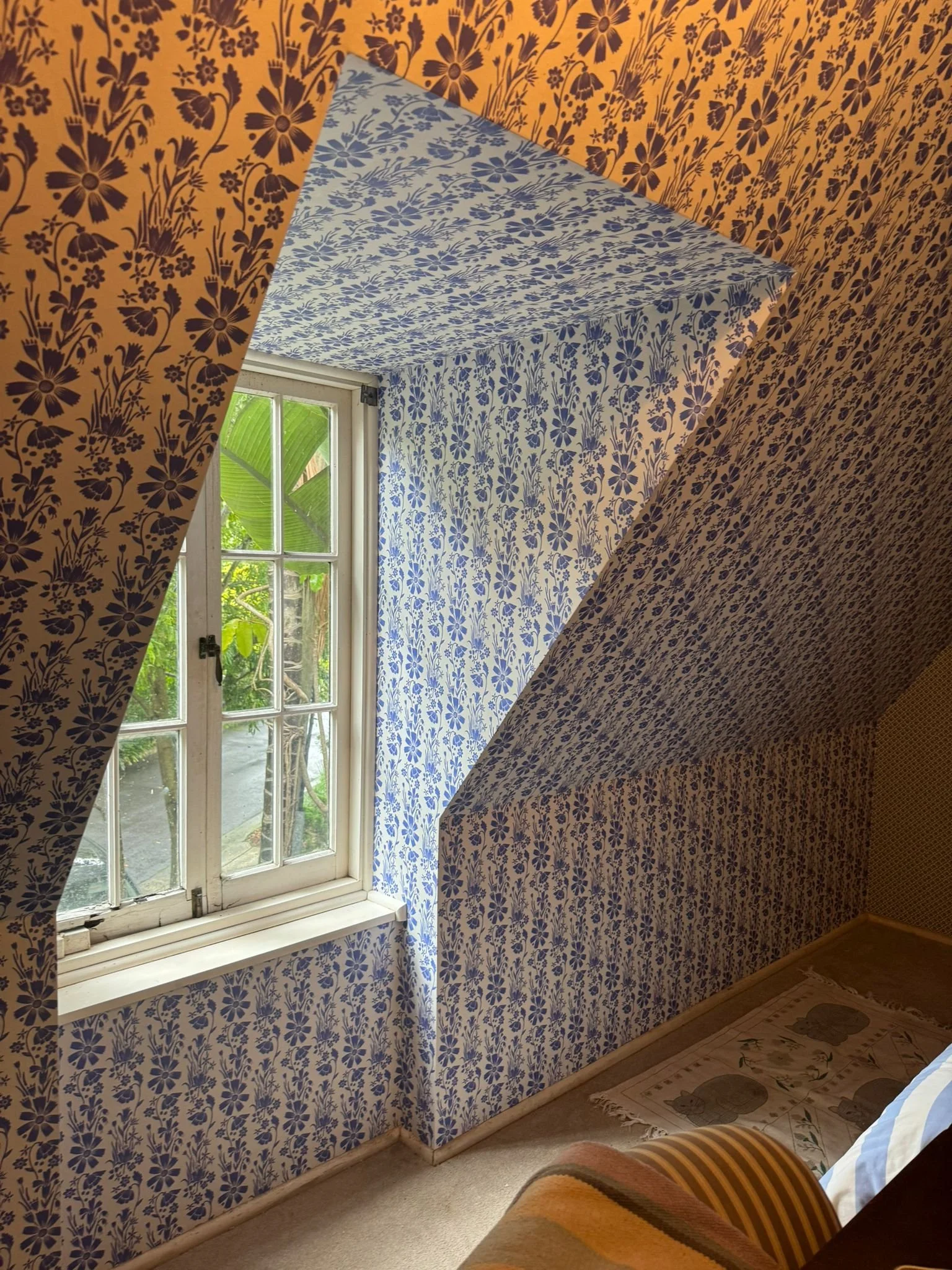A window in a room with mismatched blue and white floral wallpaper on the walls and ceiling. Outside the window, green foliage is visible. There is a small rug on the floor with an animal pattern.