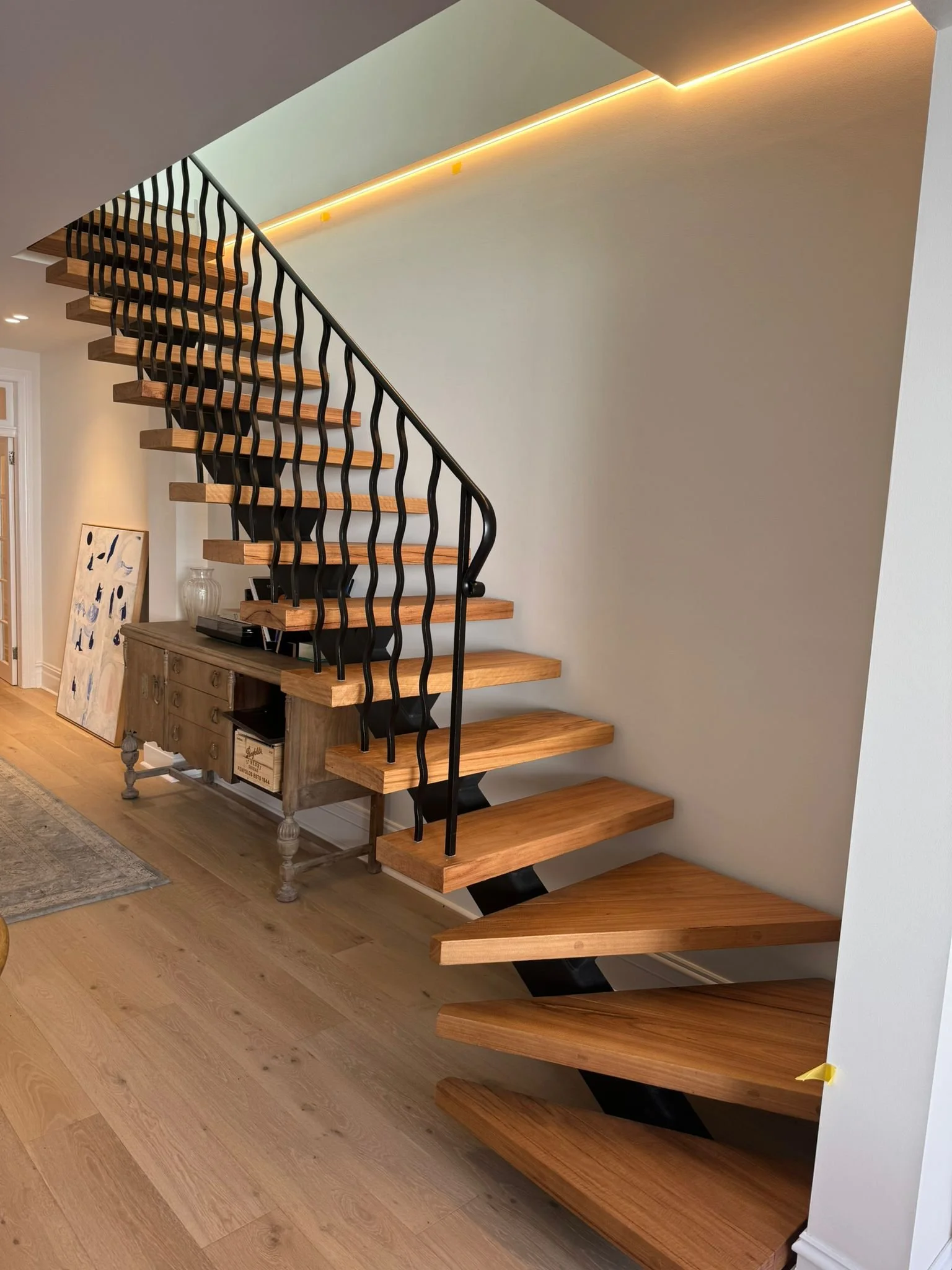 Modern wooden staircase with black metal railings, in a well-lit room with light-colored wooden flooring and a side table underneath.