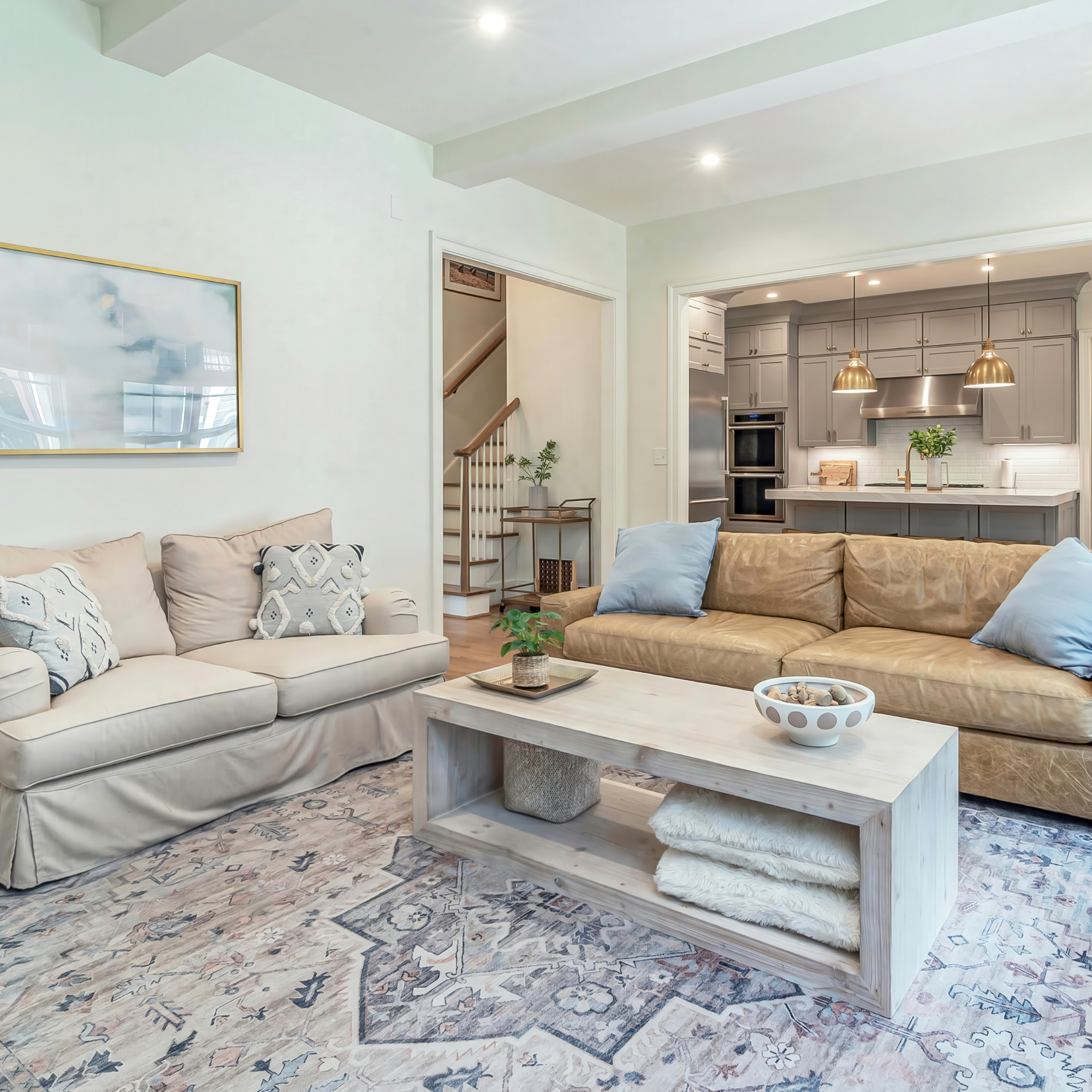 Living room with two sofas, a patterned area rug, a light wood coffee table, and view into a kitchen with gray cabinets and brass pendant lights.