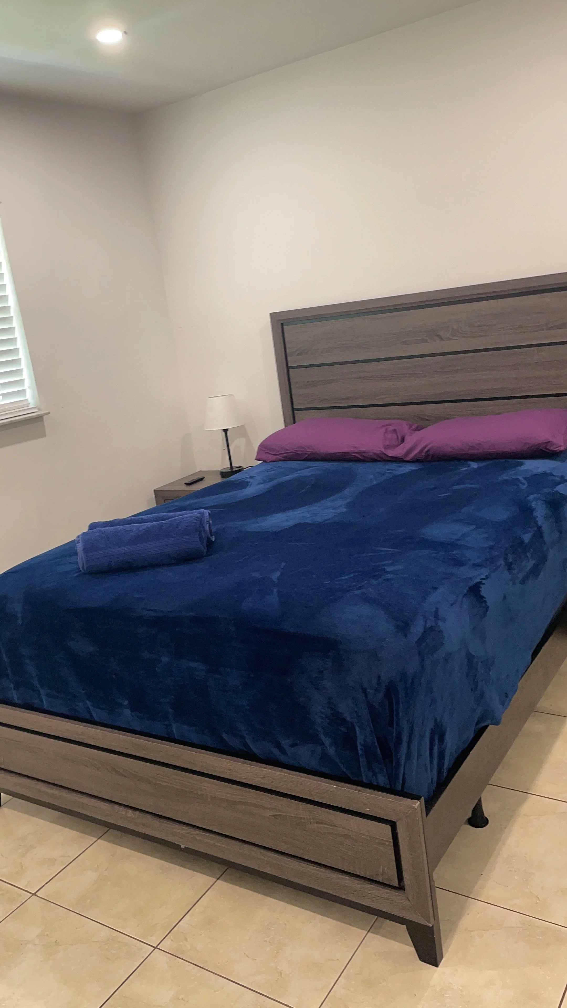 A bedroom with a wooden bed frame, purple pillow, blue blanket, and a rolled towel on top. There is a small bedside table with a lamp and remote control, and a window with blinds on the left wall.