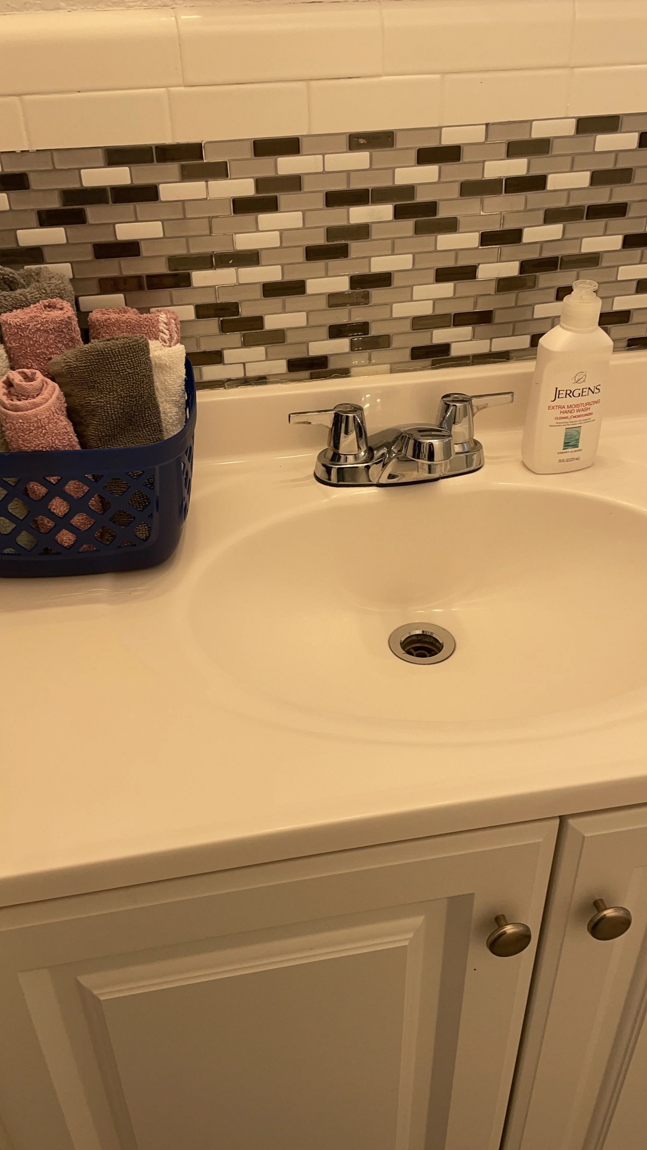 A bathroom sink with a countertop, faucet, soap dispenser, and a basket of neatly rolled towels. The backsplash features small rectangular tiles in white, gray, and black.