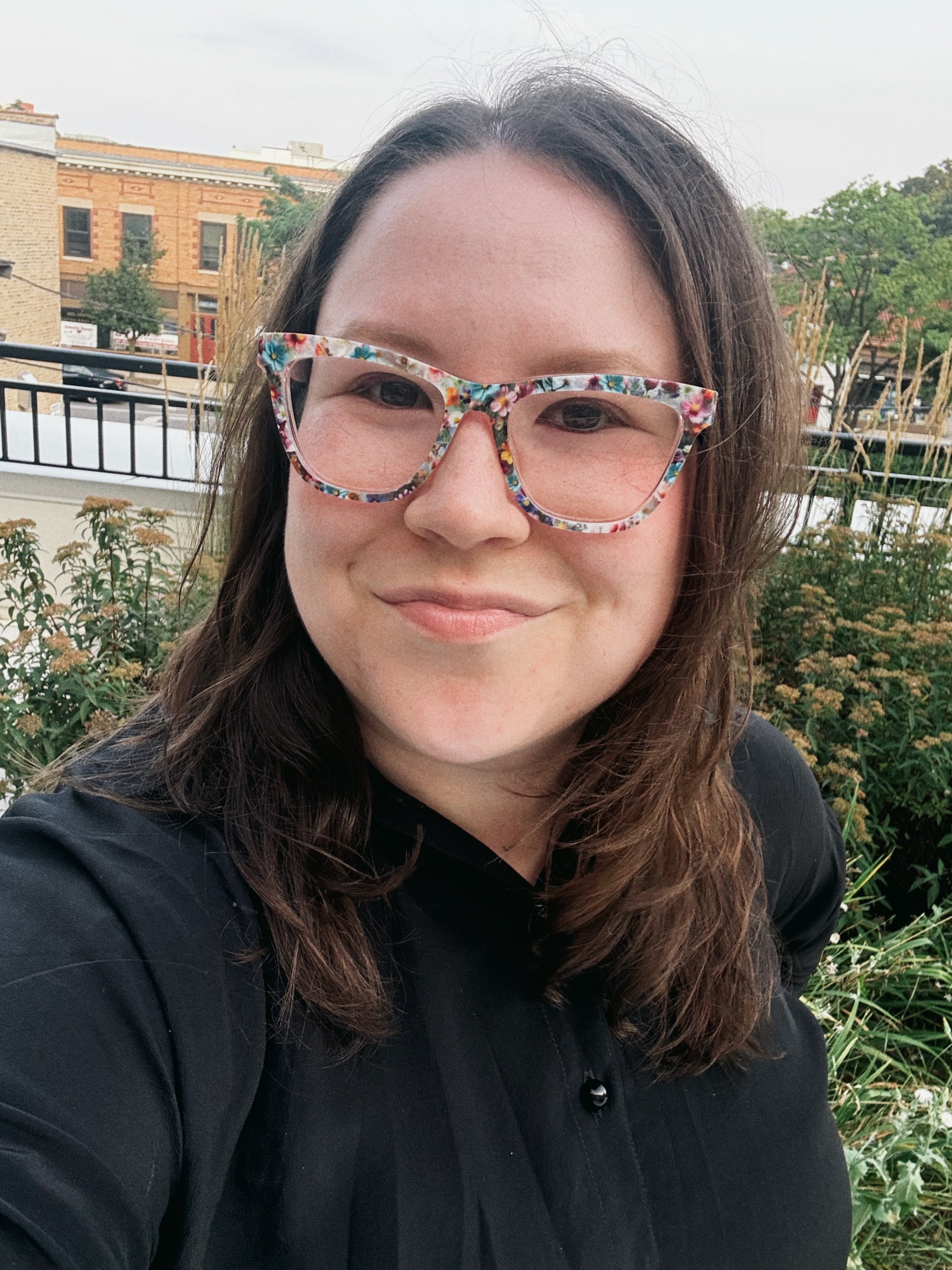 Close-up of a woman with long brown hair wearing colorful floral glasses and a black shirt, smiling outdoors with plants and buildings in the background.