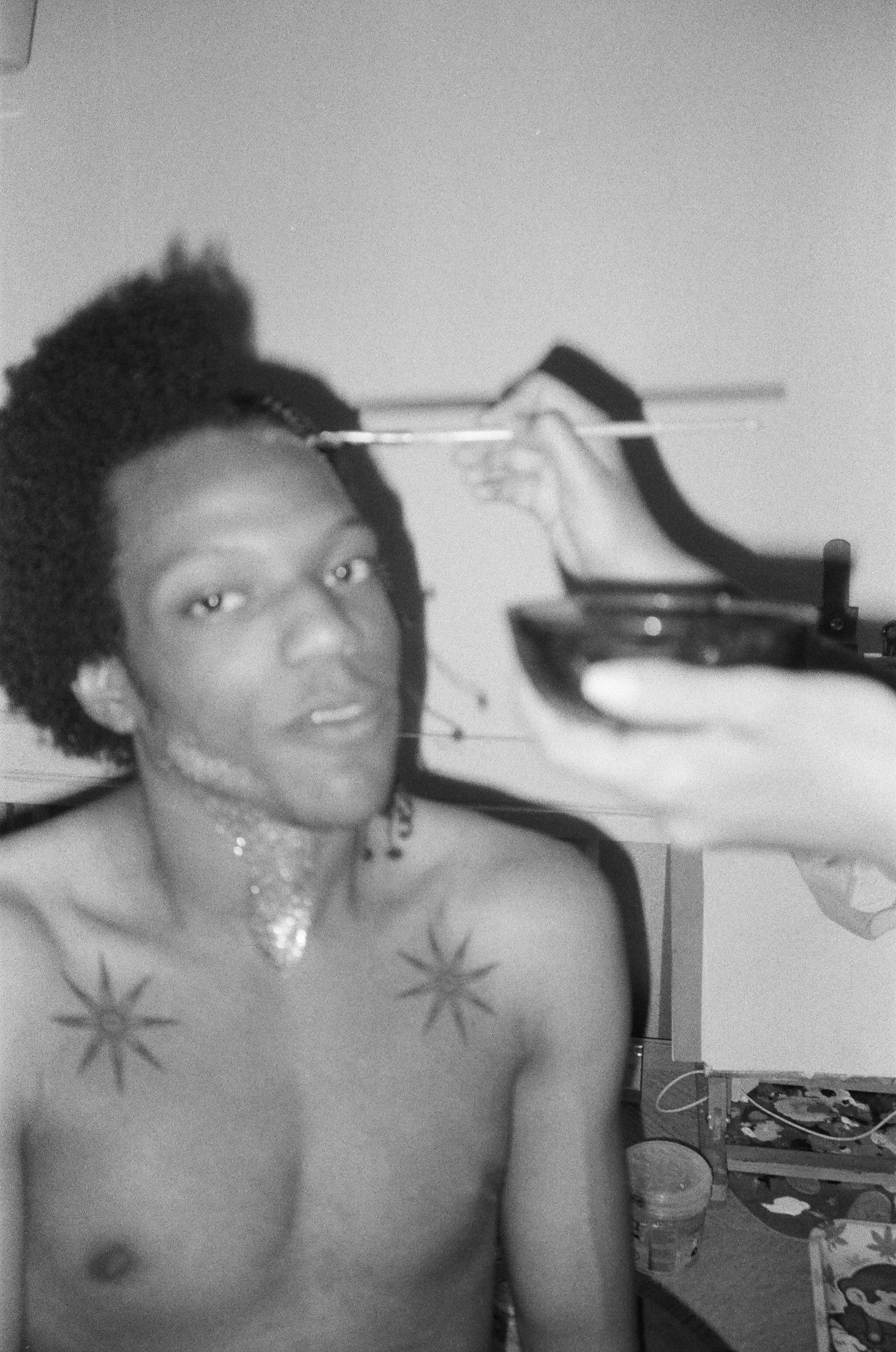 Black and white photo of a woman with freckles, curly hair, and tattoos on her chest, wearing jewelry, in a cluttered room with a laptop, a coffee mug, and various items on the table and shelves.