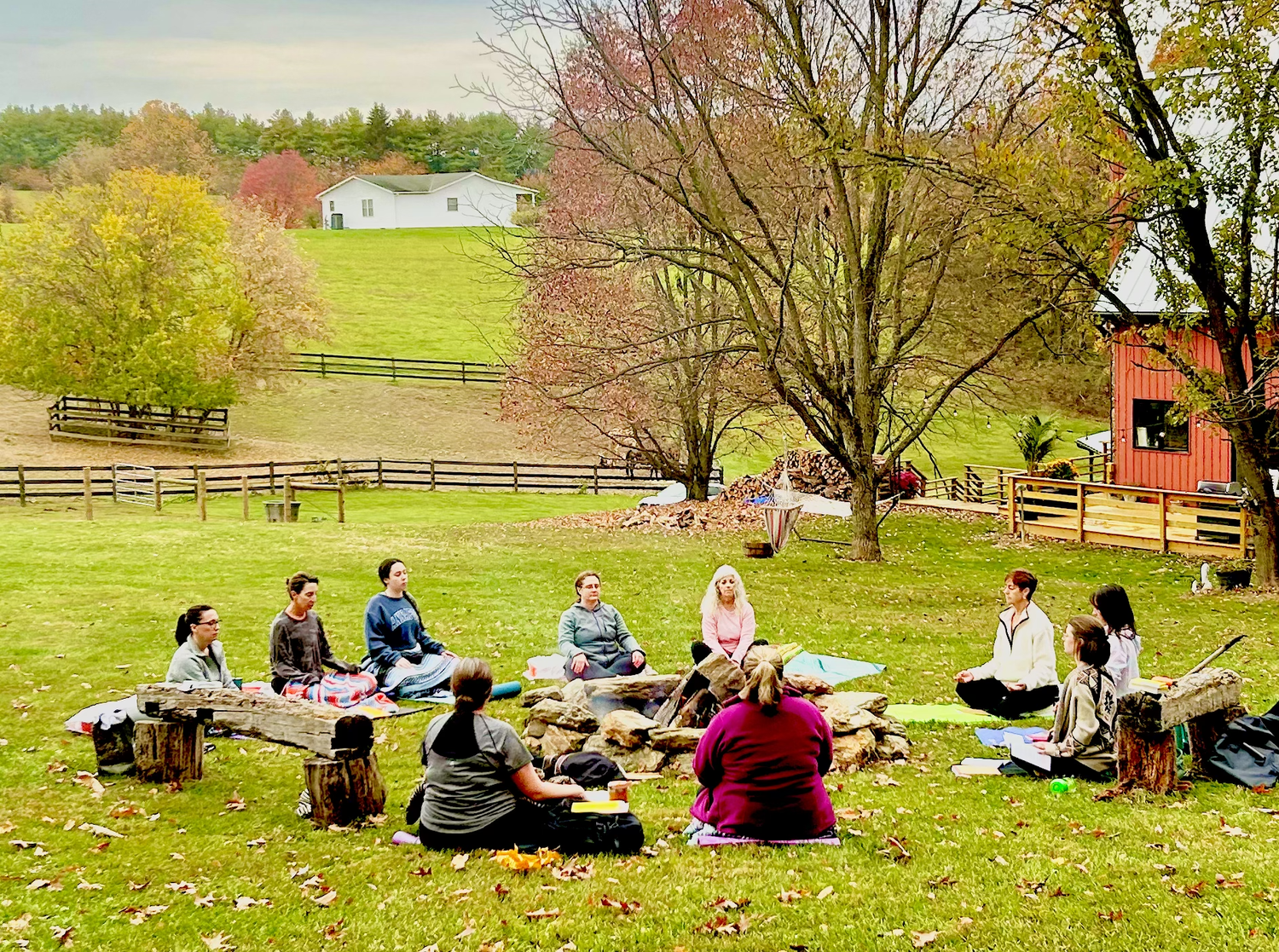 People participating in an outdoor meditation session around a fire pit in a grassy backyard with trees and houses in the background.