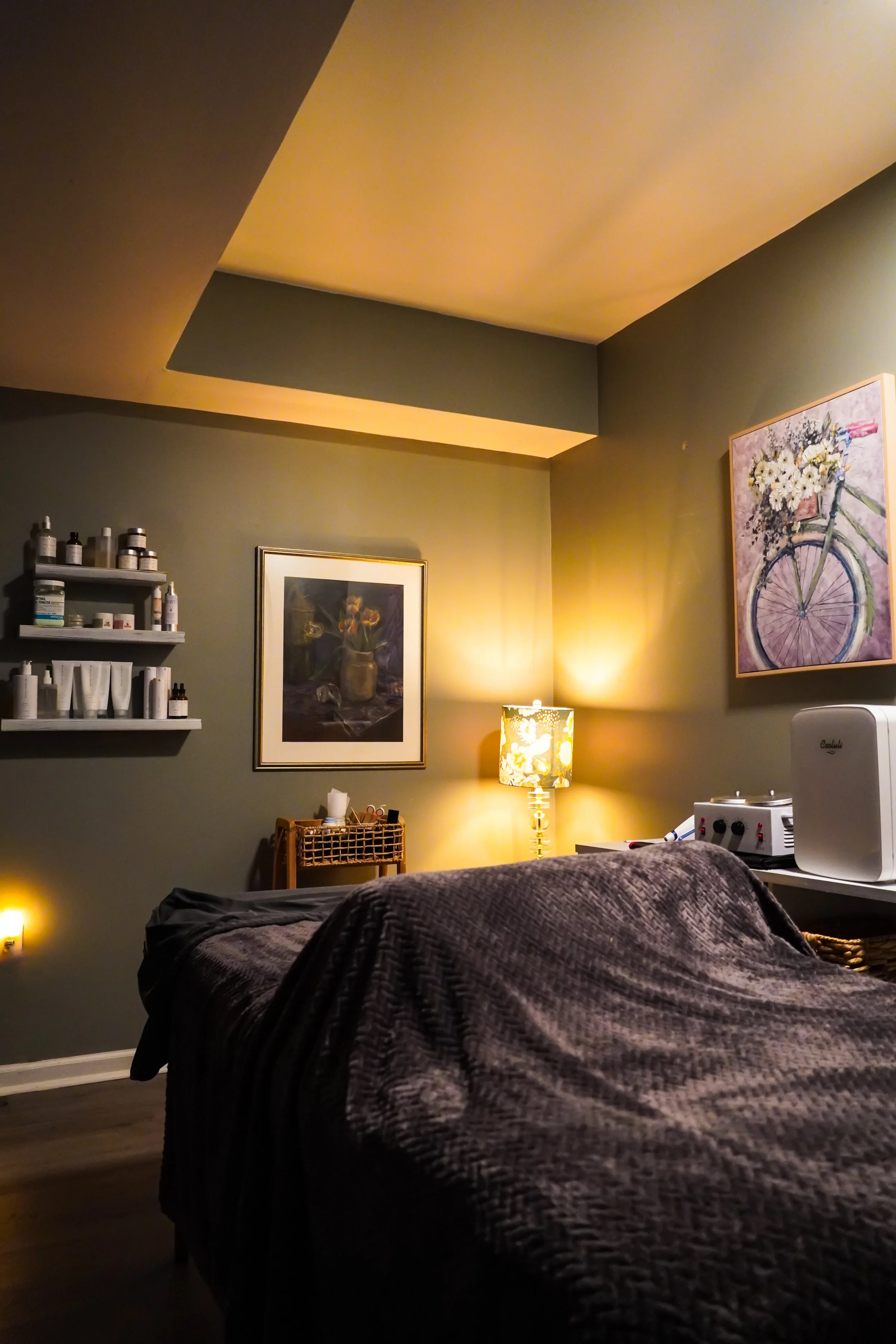 A cozy living room corner with a bed covered in a dark quilt. The wall is painted in gray and decorated with two framed pictures, one of a bicycle with flowers and another of a still life with fruit and a jug. There is a small shelf with bottles and containers, a wooden basket on the floor, and a table lamp with a floral lampshade providing warm lighting.