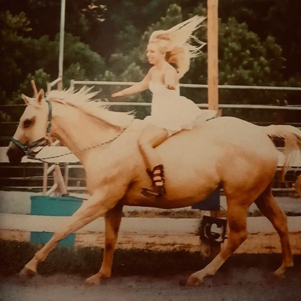 A woman with blonde hair riding a galloping tan horse in an outdoor arena, with a green background of trees and a wooden fence.