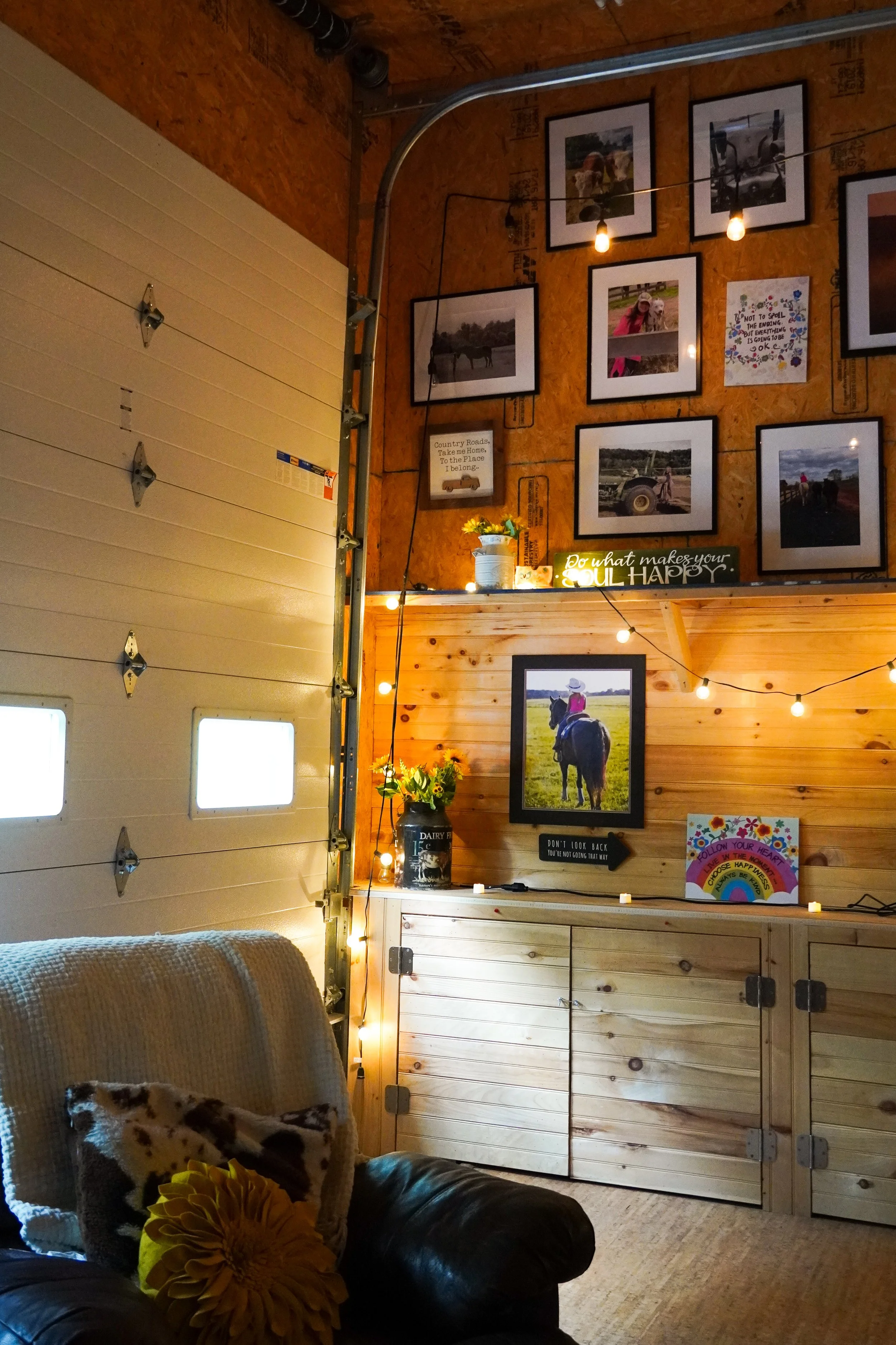 Interior of a cozy room with a animal print pillow, a bright yellow flower pillow, and a dark leather armchair in the foreground. The background features a wall with framed photos of people, horses, and outdoor scenes, along with decorative signs and string lights, and a wooden door with metal hinges and a garage-style opener.