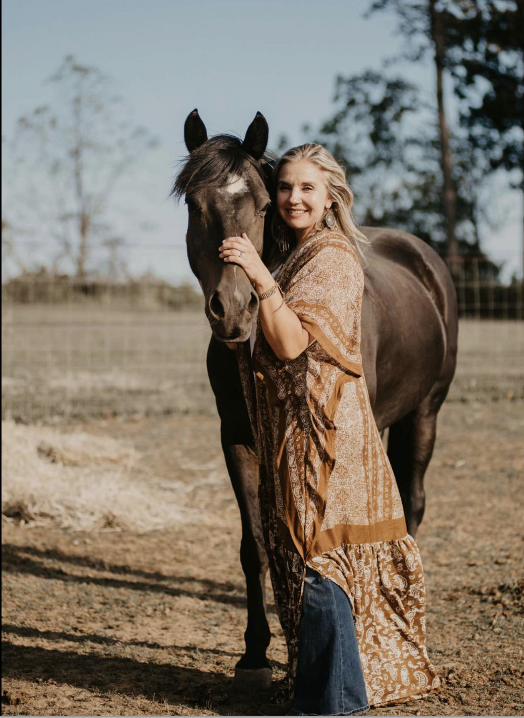 A smiling woman in a bohemian dress standing next to a dark horse, gently touching its face, on a farm with trees in the background.