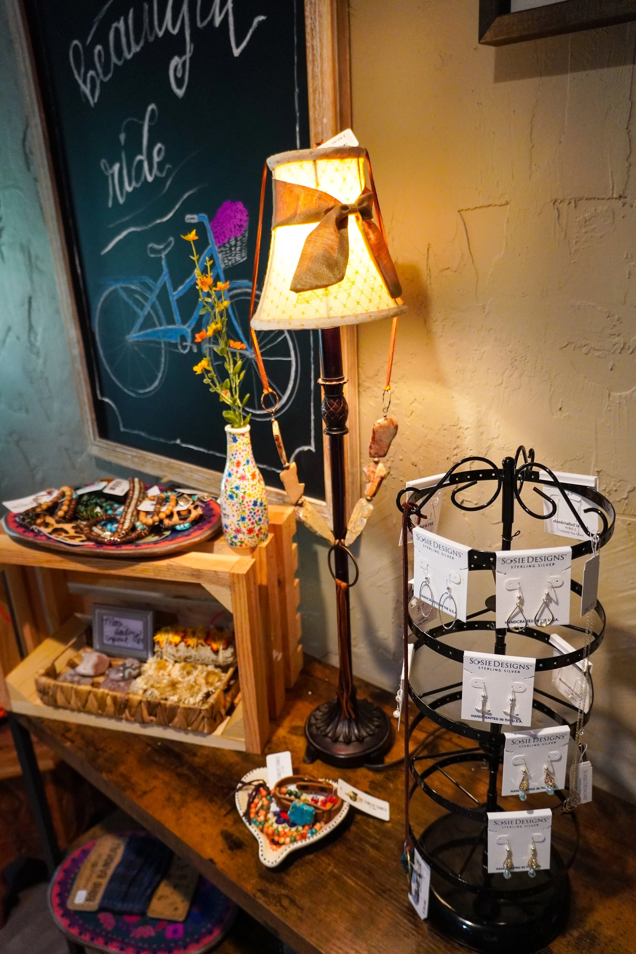 Display of jewelry, accessories, and decorative items on wooden tables and a black metal jewelry stand in a boutique or craft shop.