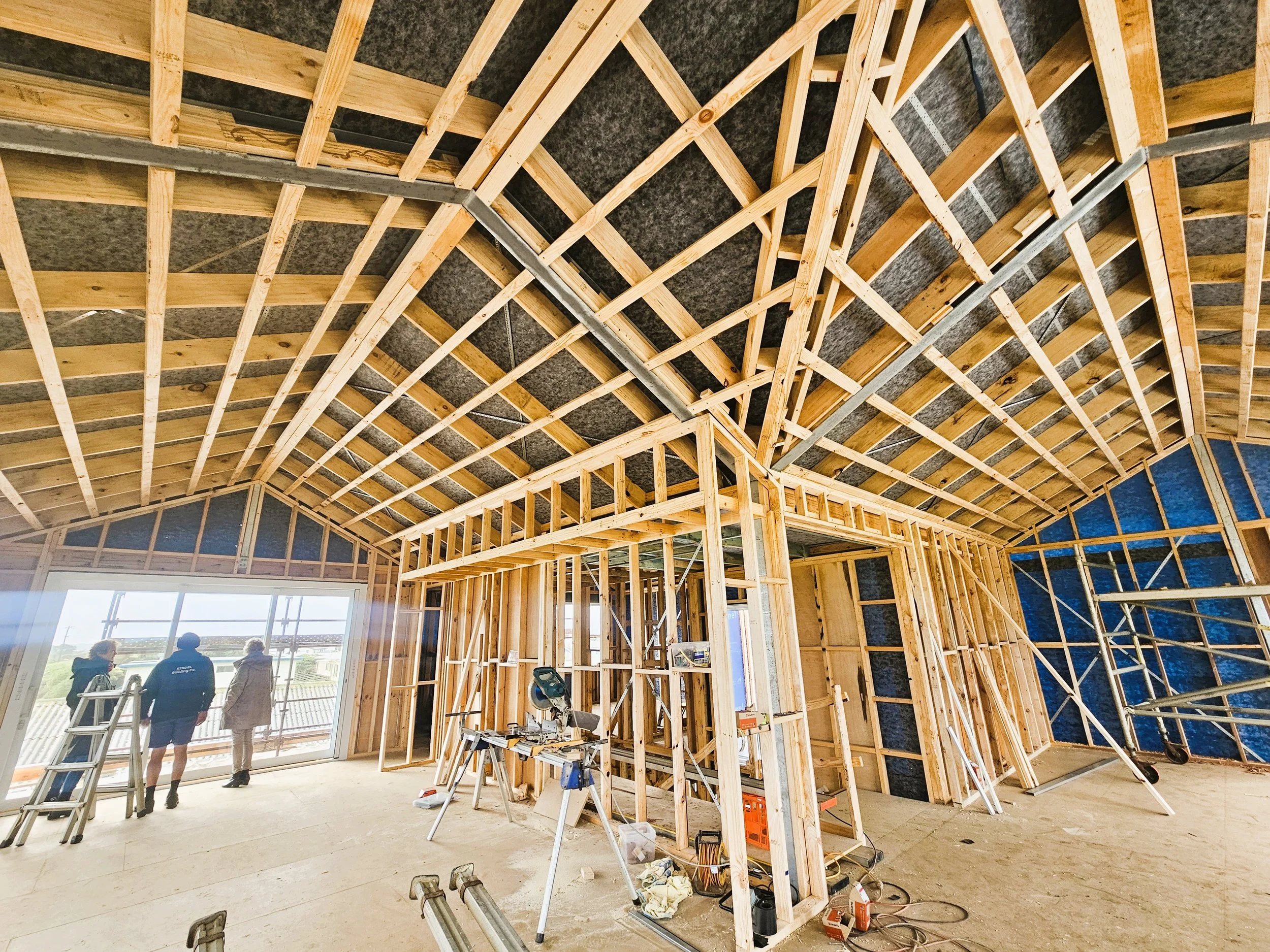 Interior view of a house under construction with exposed wooden framing and workers near a large window opening.