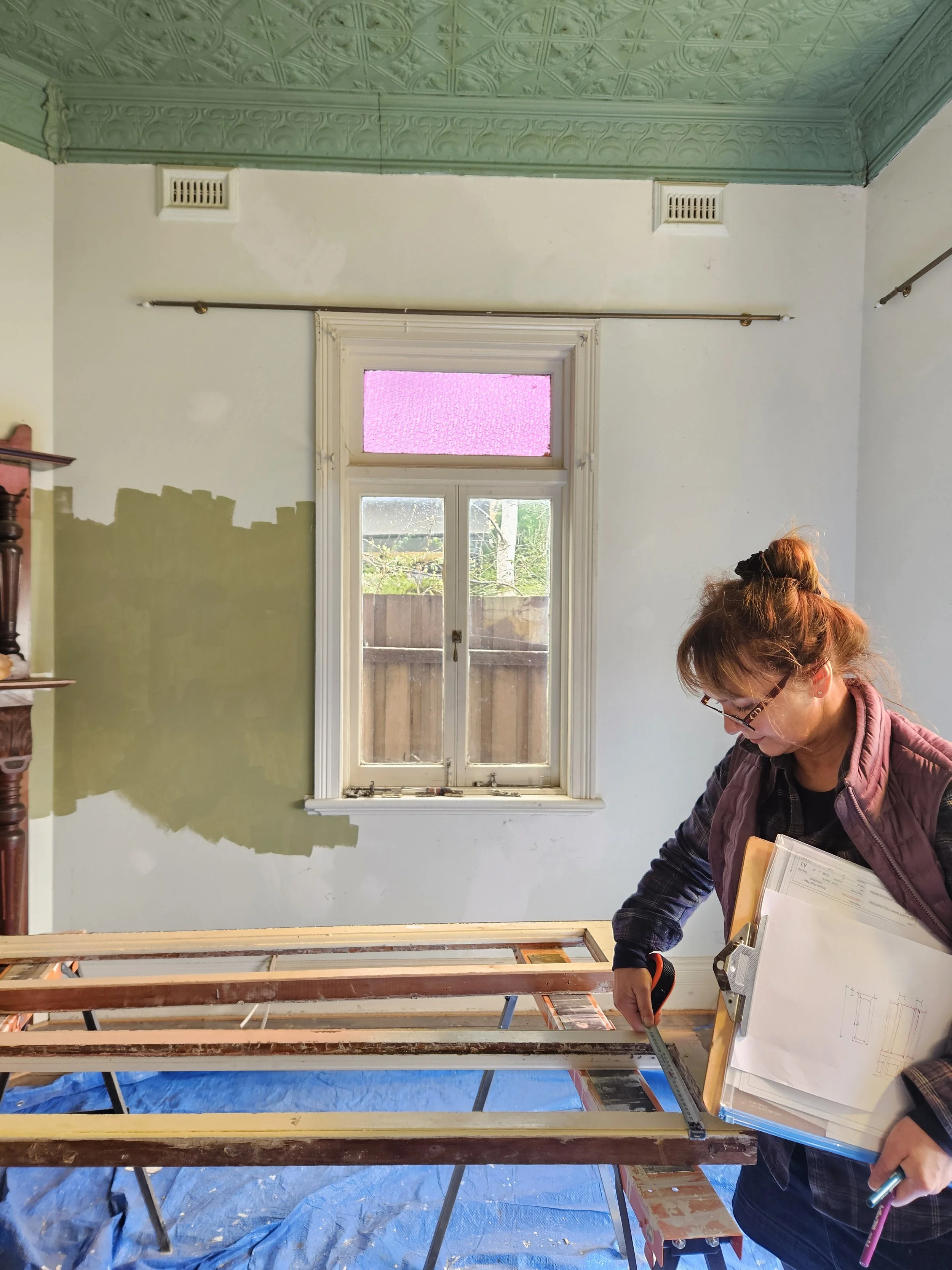 A woman is measuring a doorway frame during a home renovation inside a room with a window and an ornate ceiling. The room is partially painted and there is a blue tarp on the floor.