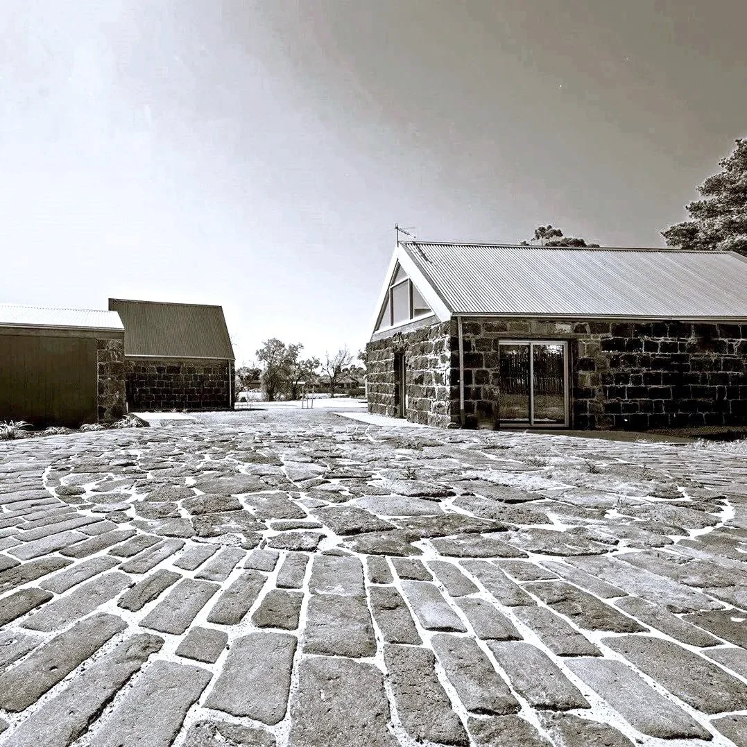 A cobblestone pathway leading to rustic stone buildings with metal roofs, set in a snowy landscape under an overcast sky.