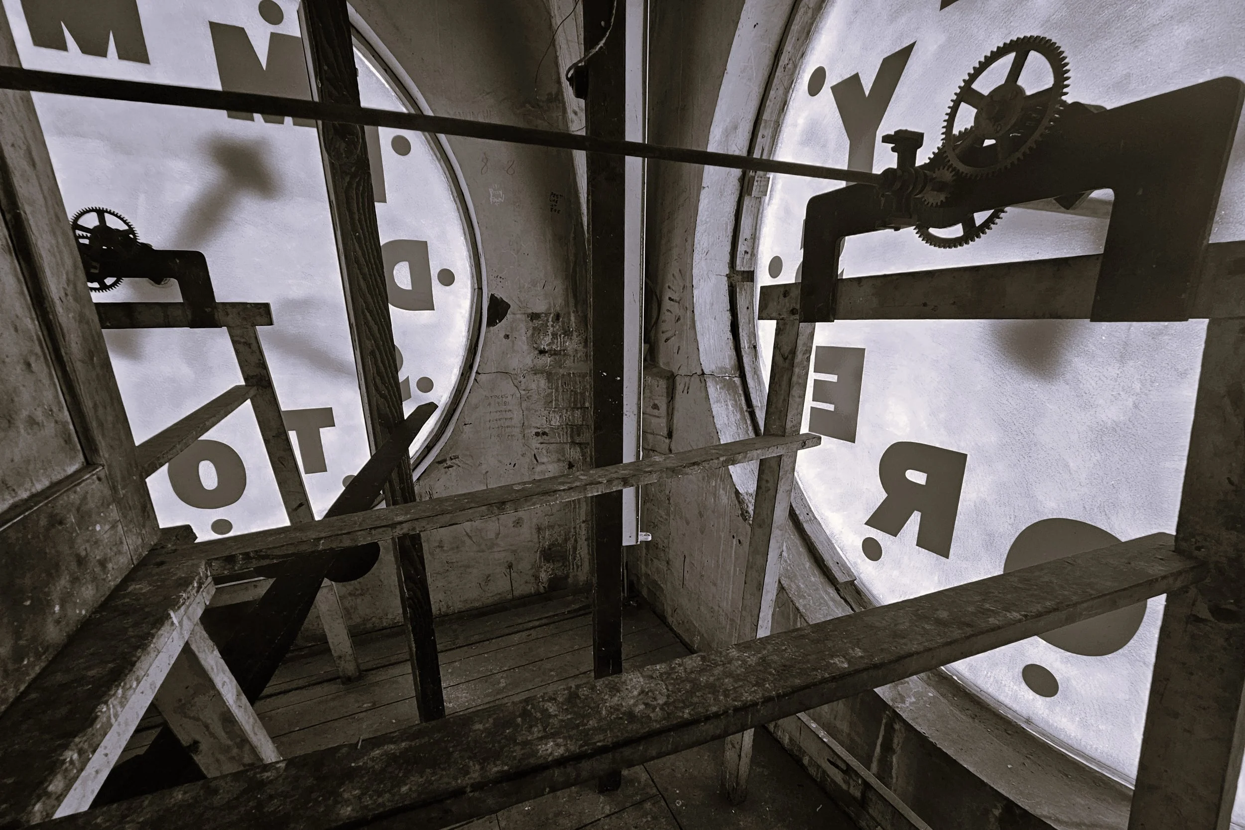 View from inside a clock tower showing the clock face and its metal gears and mechanisms, with wooden beams and scaffolding.