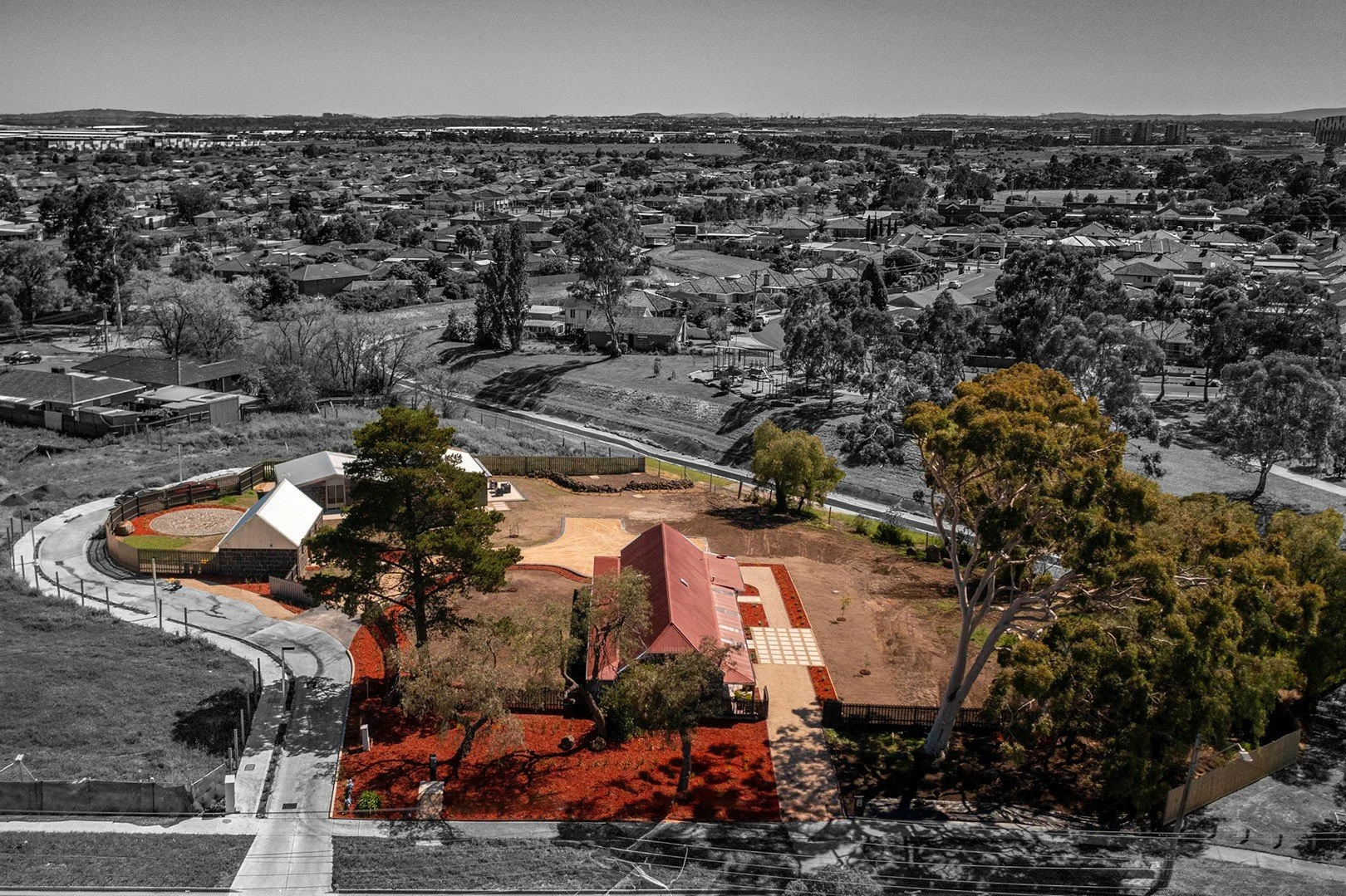 Aerial view of a park with a central building, trees, and pathways, in a residential area with houses and streets in the background.