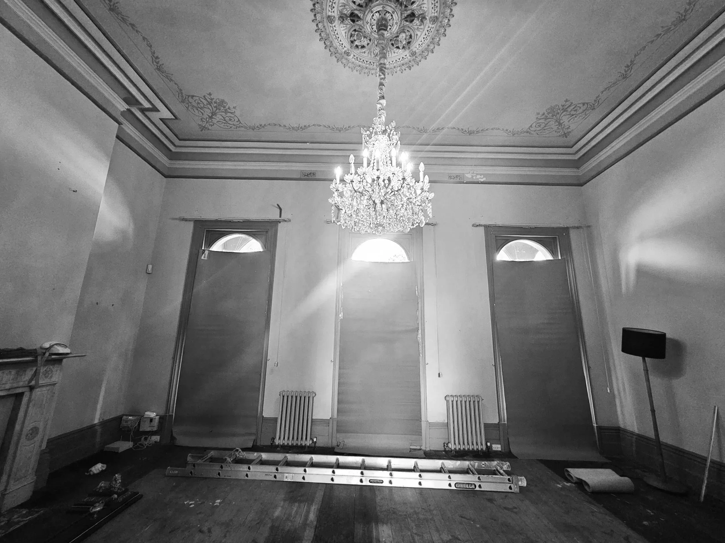 An ornate chandelier hangs from a decorated ceiling in a large room with three tall windows. Construction tools, a ladder, and a rolled-up carpet are on the floor, indicating ongoing renovation.