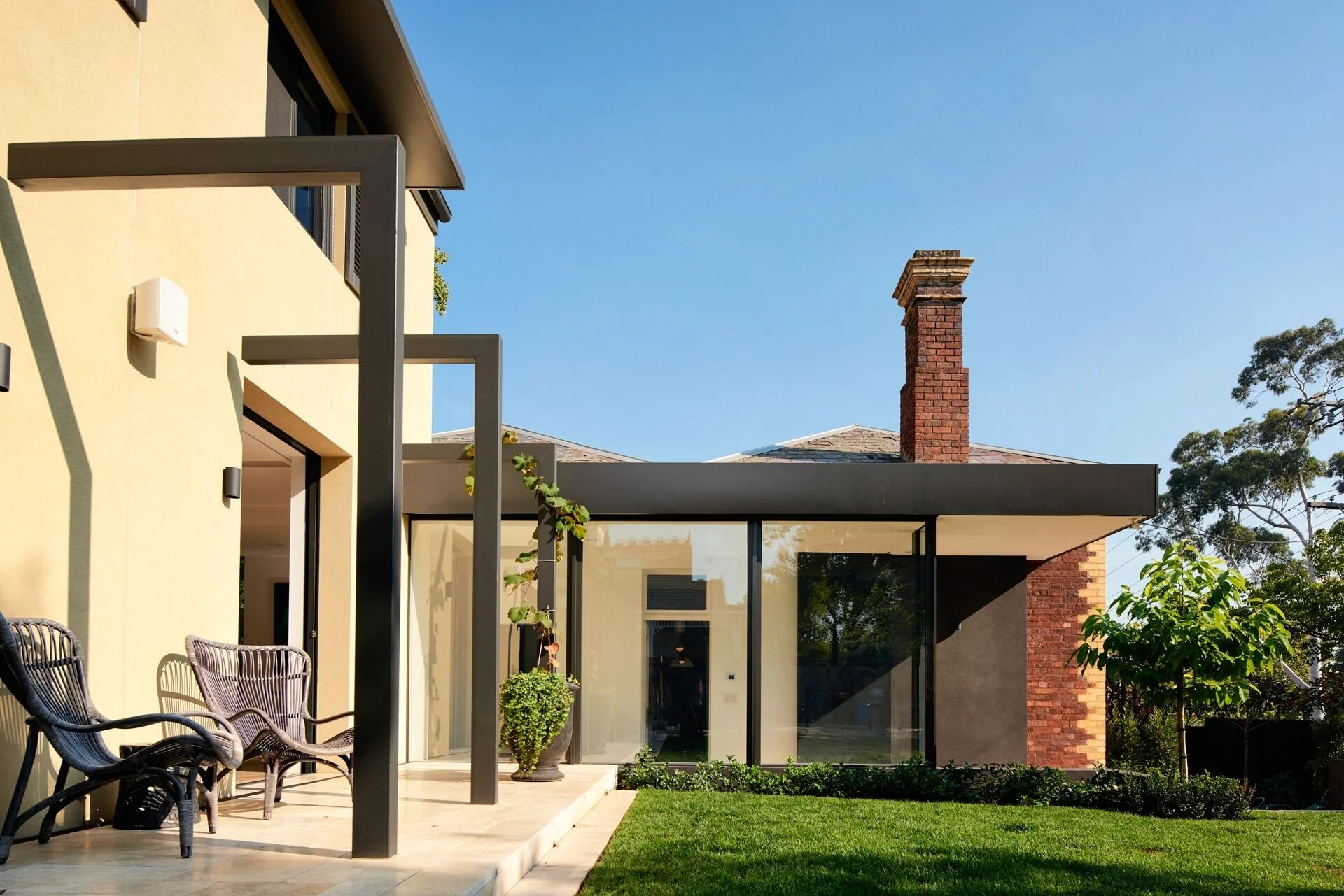 Modern house with a glass addition, yellow exterior wall, outdoor seating area with black chairs, green plants, and a red brick chimney against a clear blue sky.
