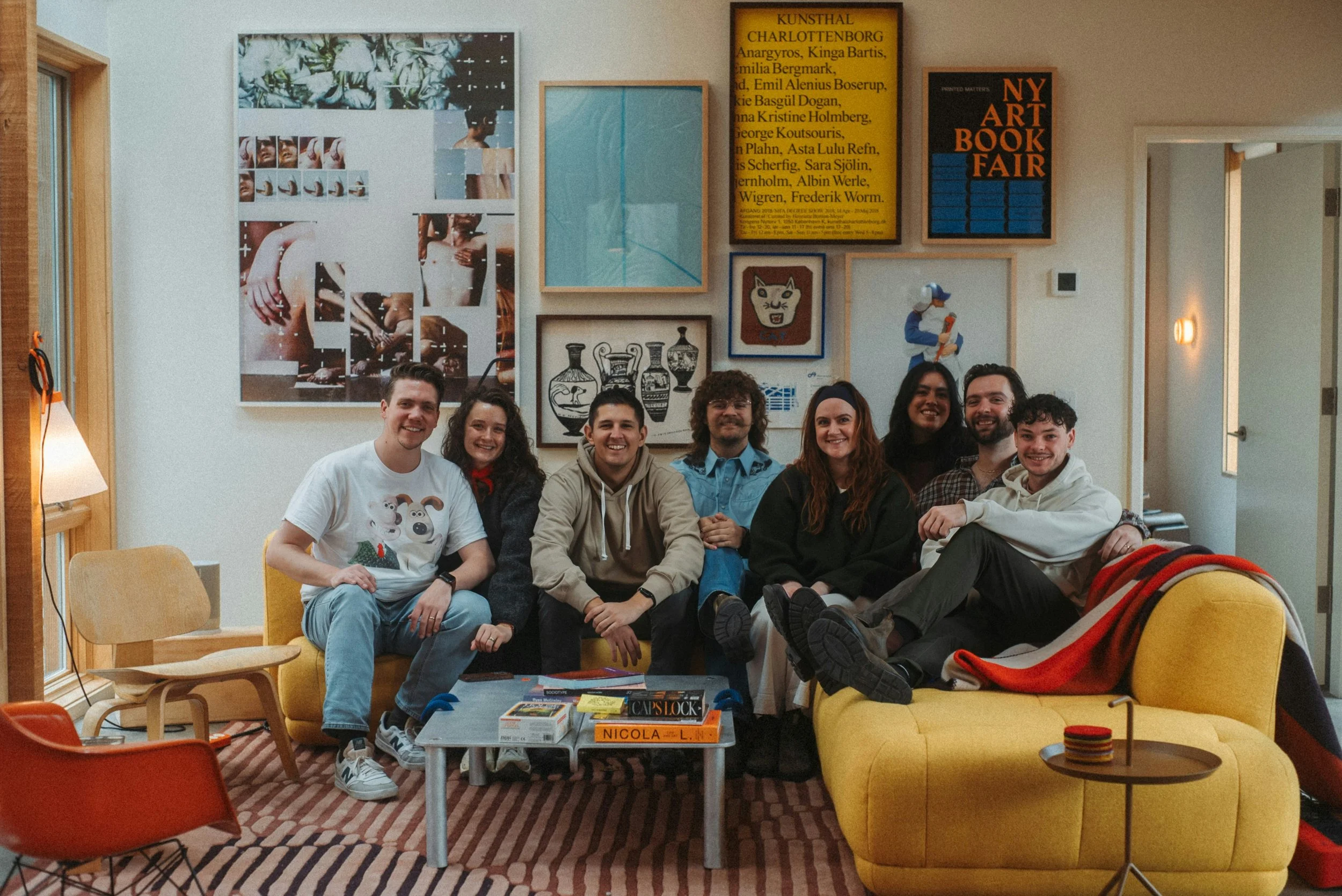 Group of seven people sitting on a yellow couch and a yellow chair in a room decorated with framed artwork and posters on the wall, with books and board games on a coffee table in front of them.