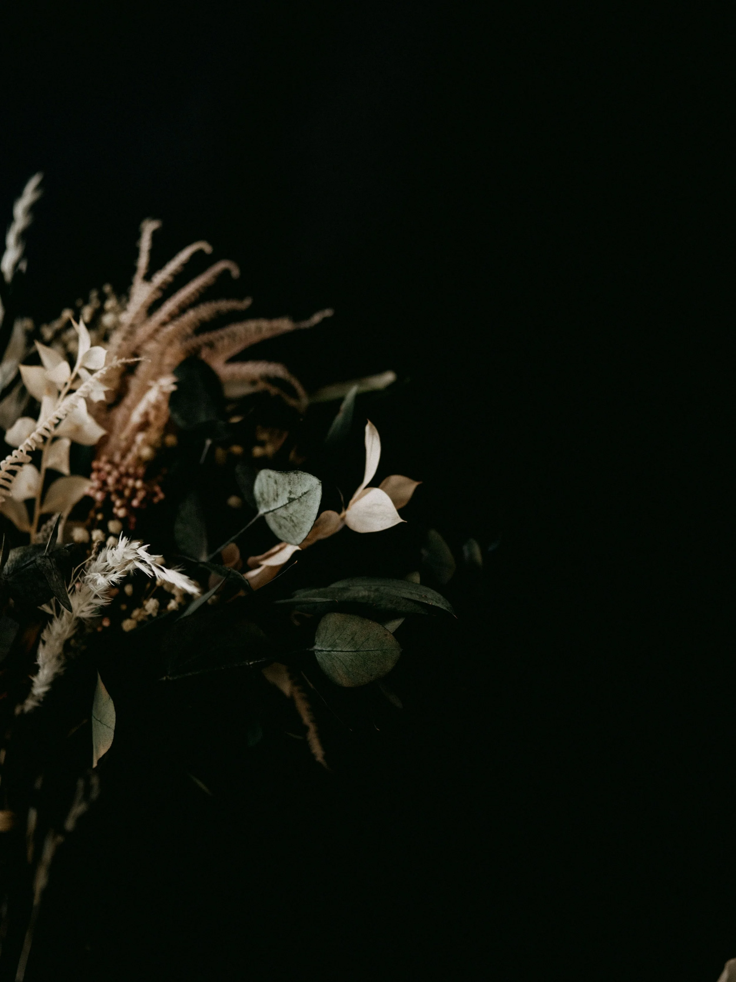 A floral arrangement with white and beige flowers and green foliage on a dark background.