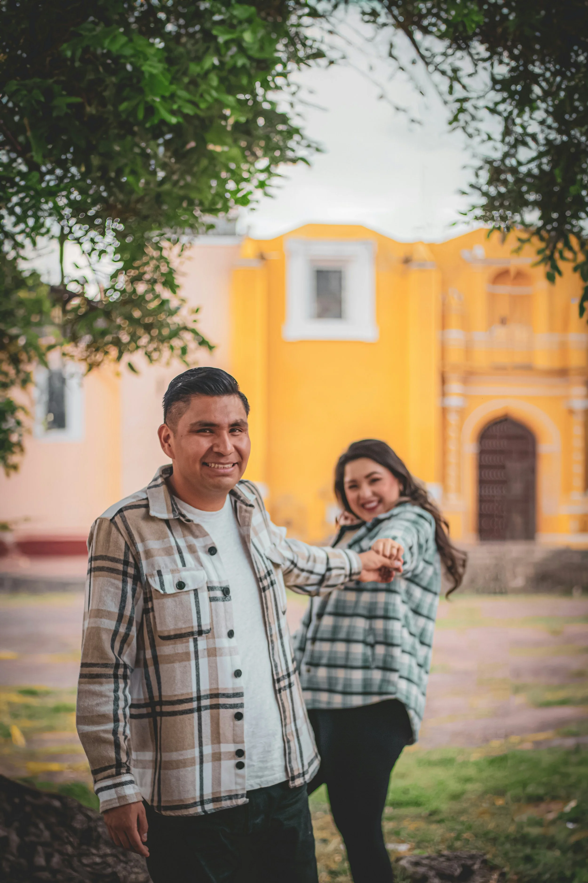 A smiling man in a plaid shirt and a woman in a plaid shirt holding hands outdoors in front of a yellow building with trees around.