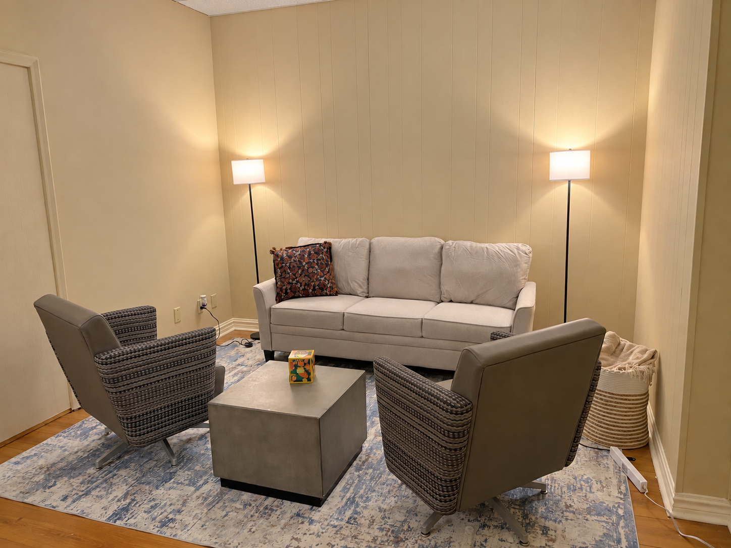 Living room with three tall floor lamps, a white sofa with a patterned pillow, two gray and black patterned armchairs, a square coffee table with a colorful toy on it, a blue and beige patterned area rug, and a cream-colored woven basket in the corner.
