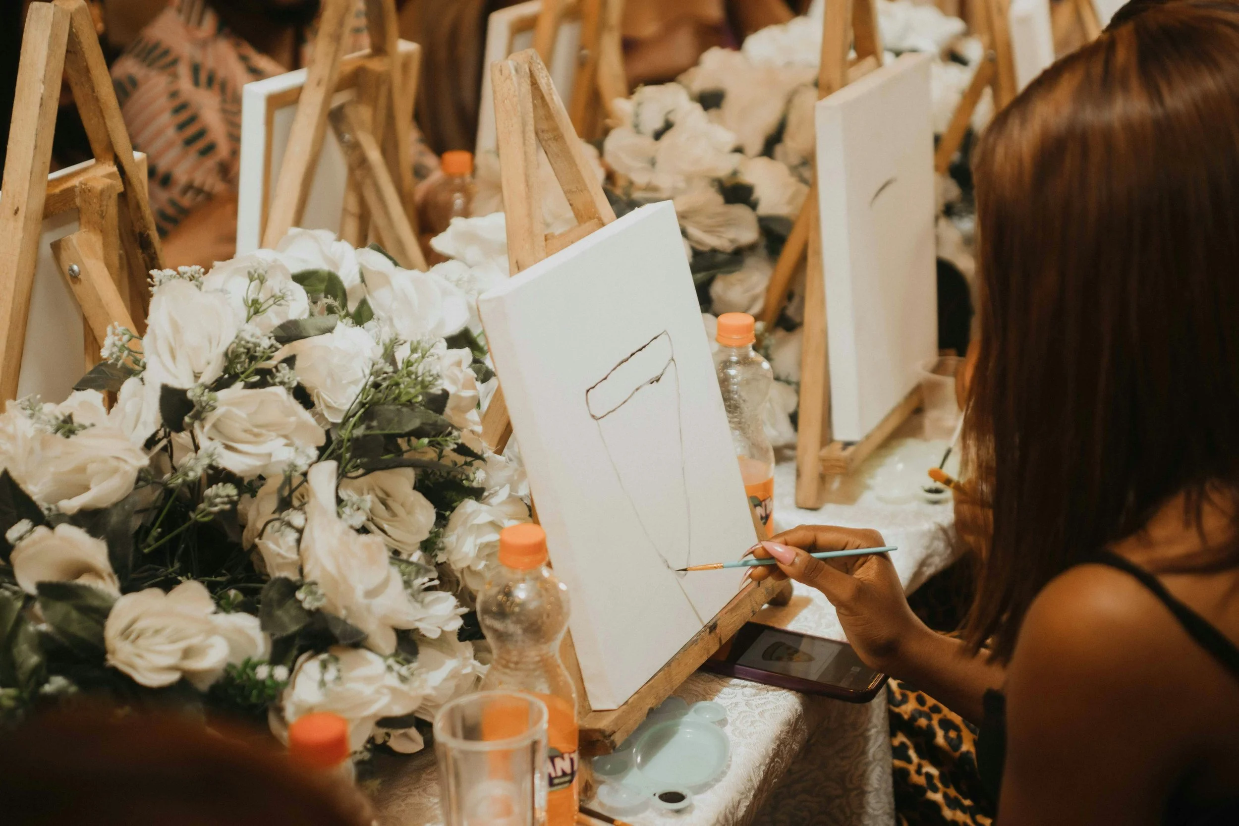 A woman painting on a canvas surrounded by white flowers and art supplies in a studio or workshop setting.