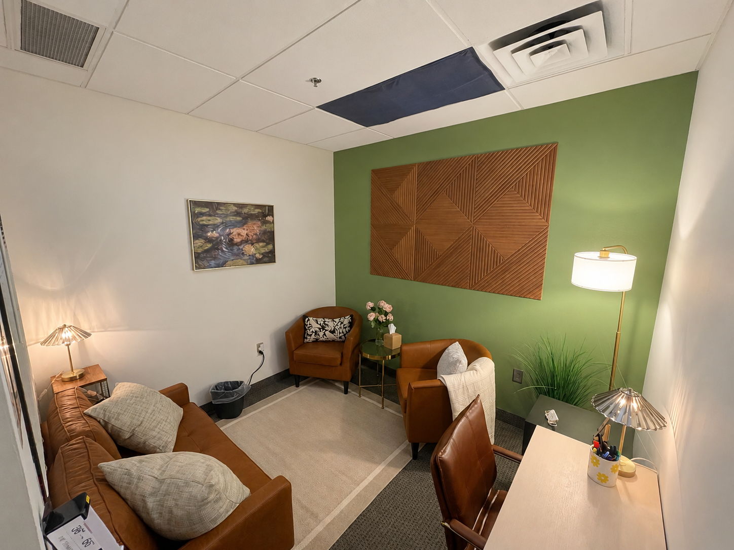 Small office lounge with a green wall, leather sofa, two armchairs, and a small table with flowers. There's a desk with a lamp and stationery, painting on the wall, and overhead ceiling tiles.