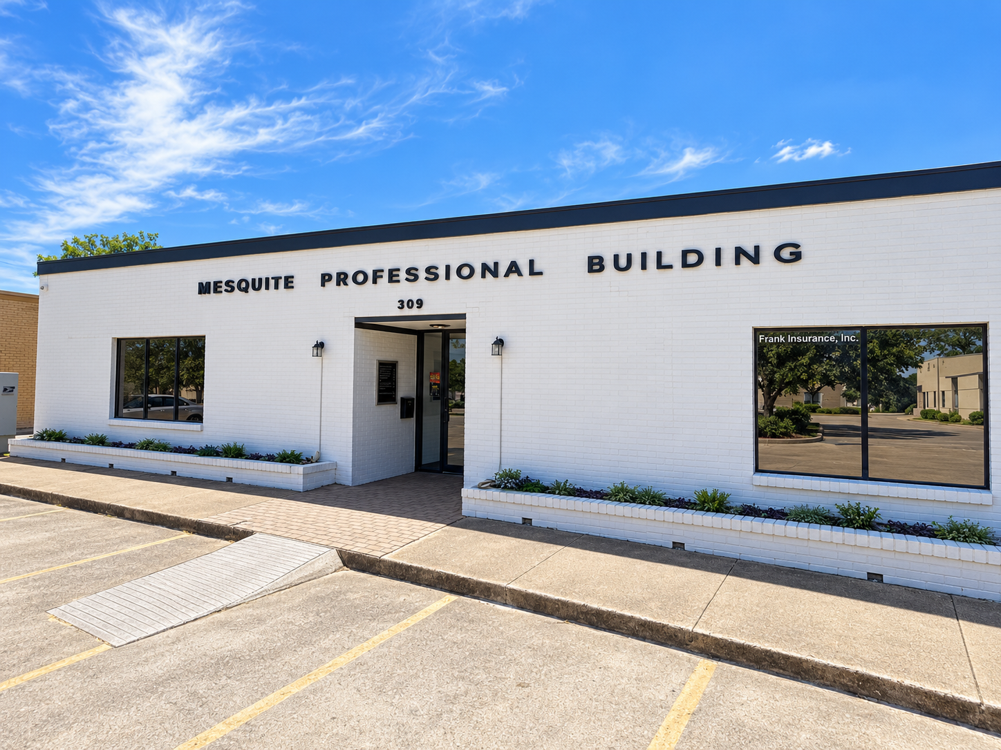 A white brick building with black lettering reads 'Mesquite Professional Building' at 309. There are large windows, some reflecting trees and neighboring buildings, and a small landscaped area with plants. The sky is blue with some clouds.