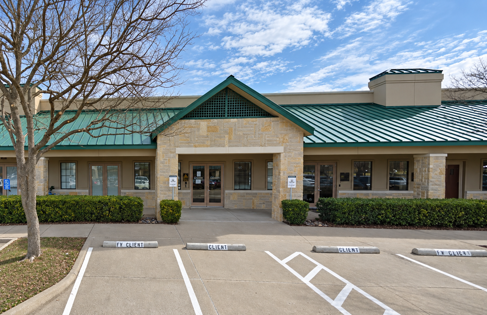 A beige building with a green metal roof and stone accents, surrounded by a parking lot with designated spaces marked "CLIENT" and "FK CLIENT." There are two signs in front of the building that read "Client." A leafless tree is on the left side, and a clear sky with some clouds is above.