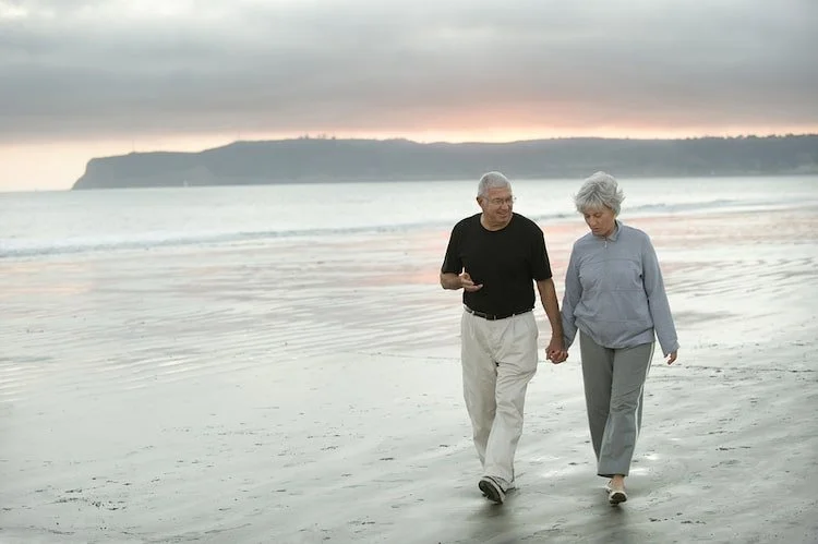 An elderly couple walking hand in hand on the beach during sunset.