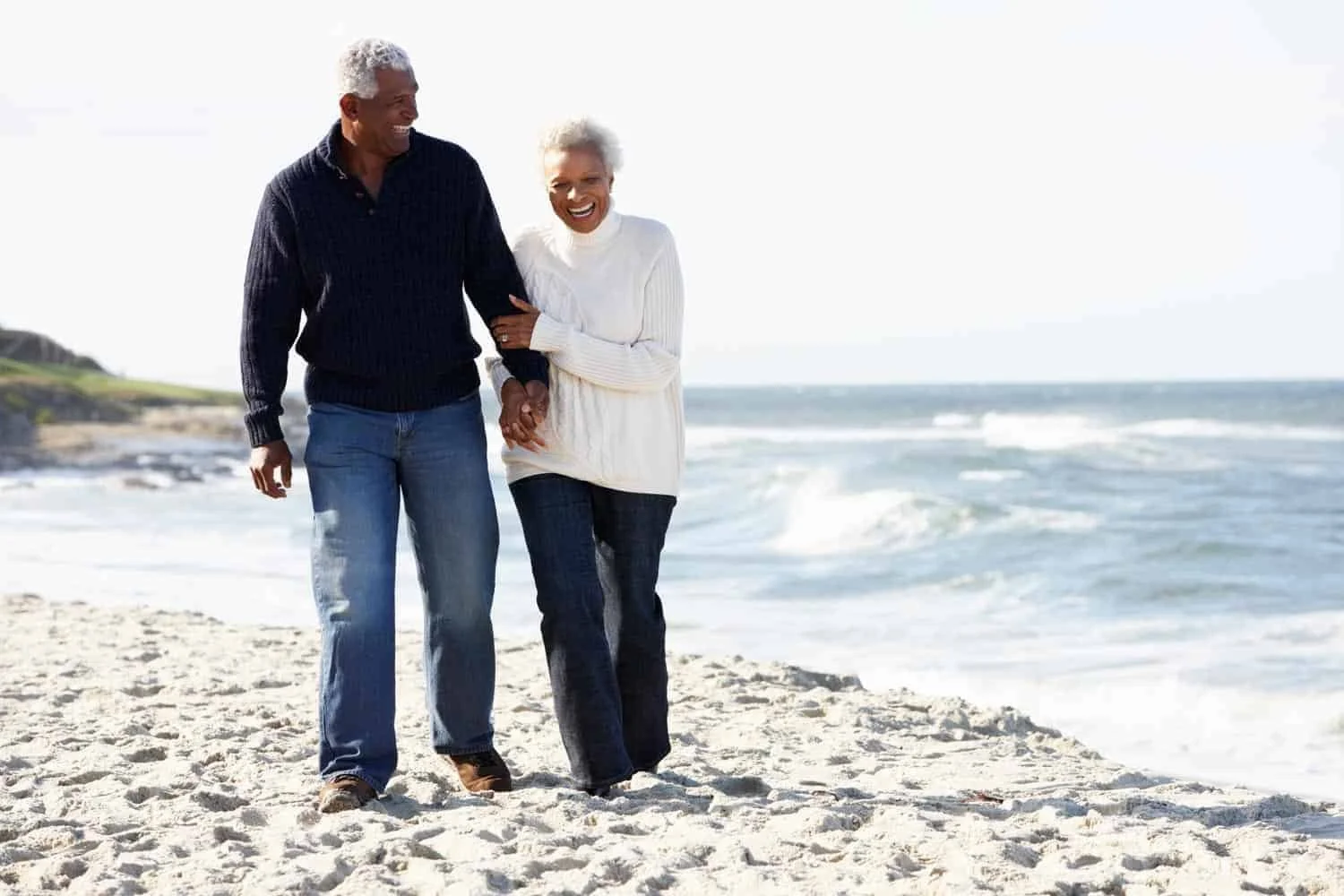 An elderly couple walking together on a sandy beach, smiling and holding hands, with the ocean waves and a clear sky in the background.