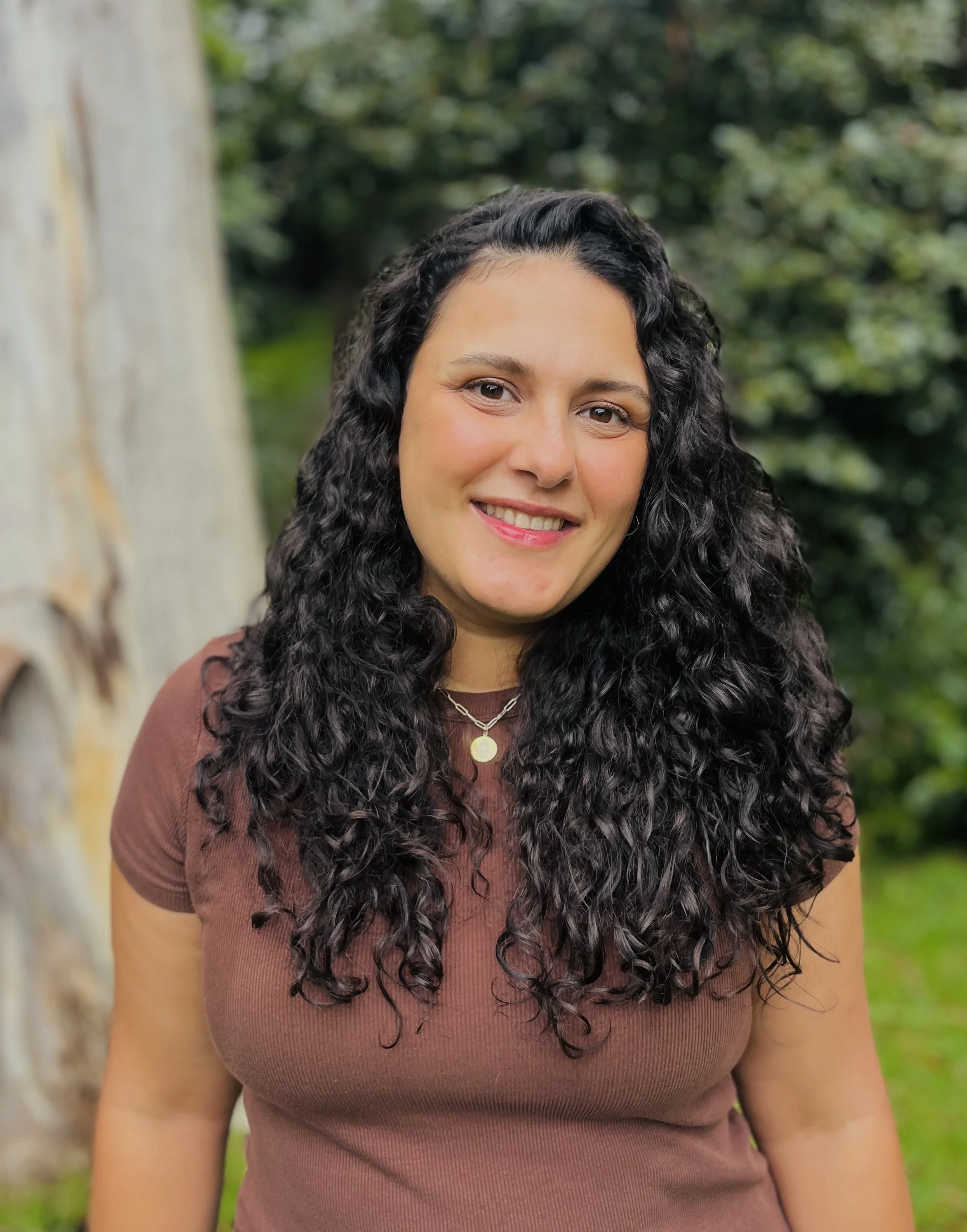 Close-up of a smiling woman with curly dark hair wearing a brown top and a gold necklace, standing outdoors with blurred greenery and a large tree in the background.