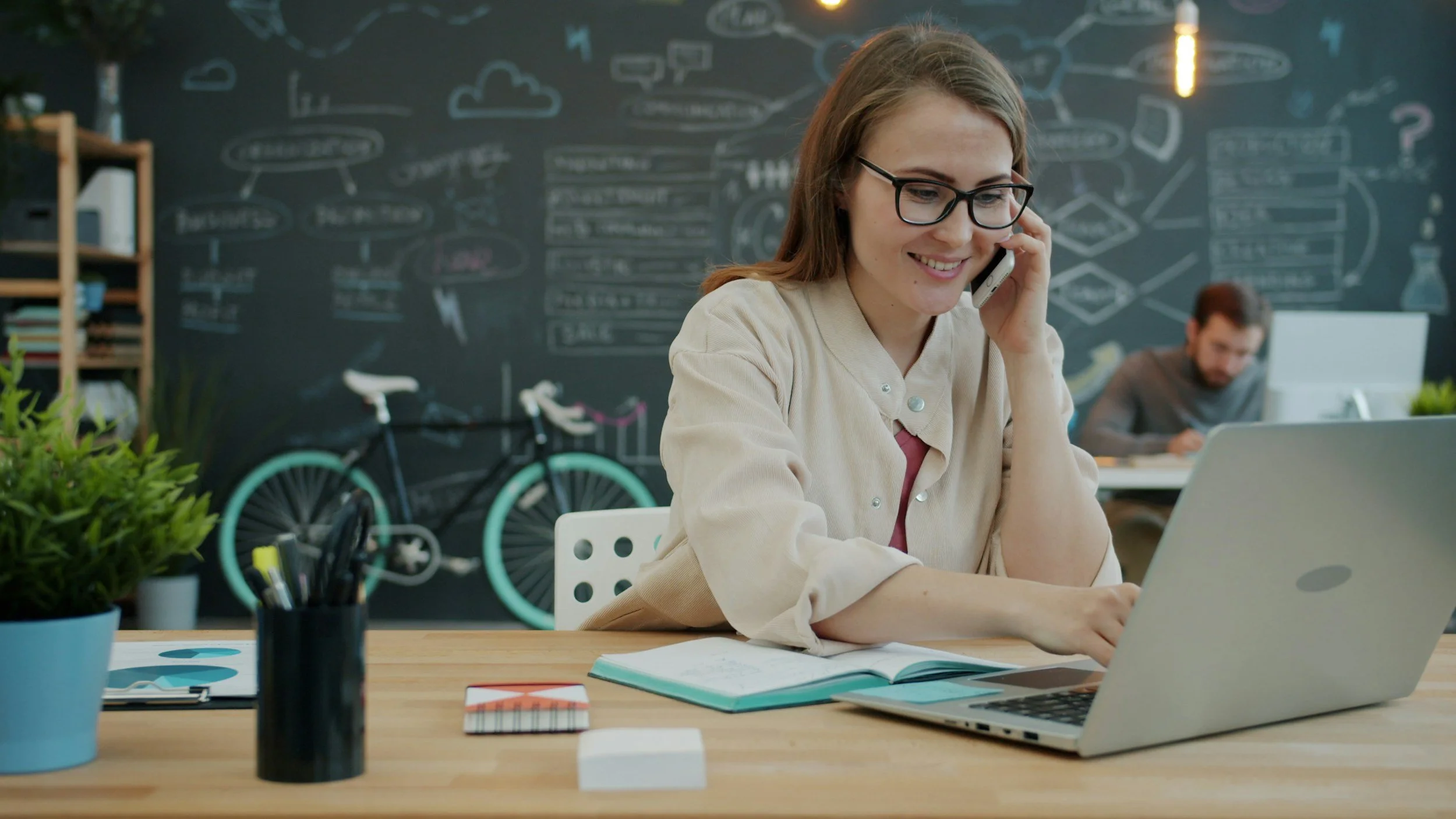 A woman with glasses talking on a cell phone while working on a laptop at a desk in a modern office with a dark chalkboard wall decorated with diagrams and drawings.