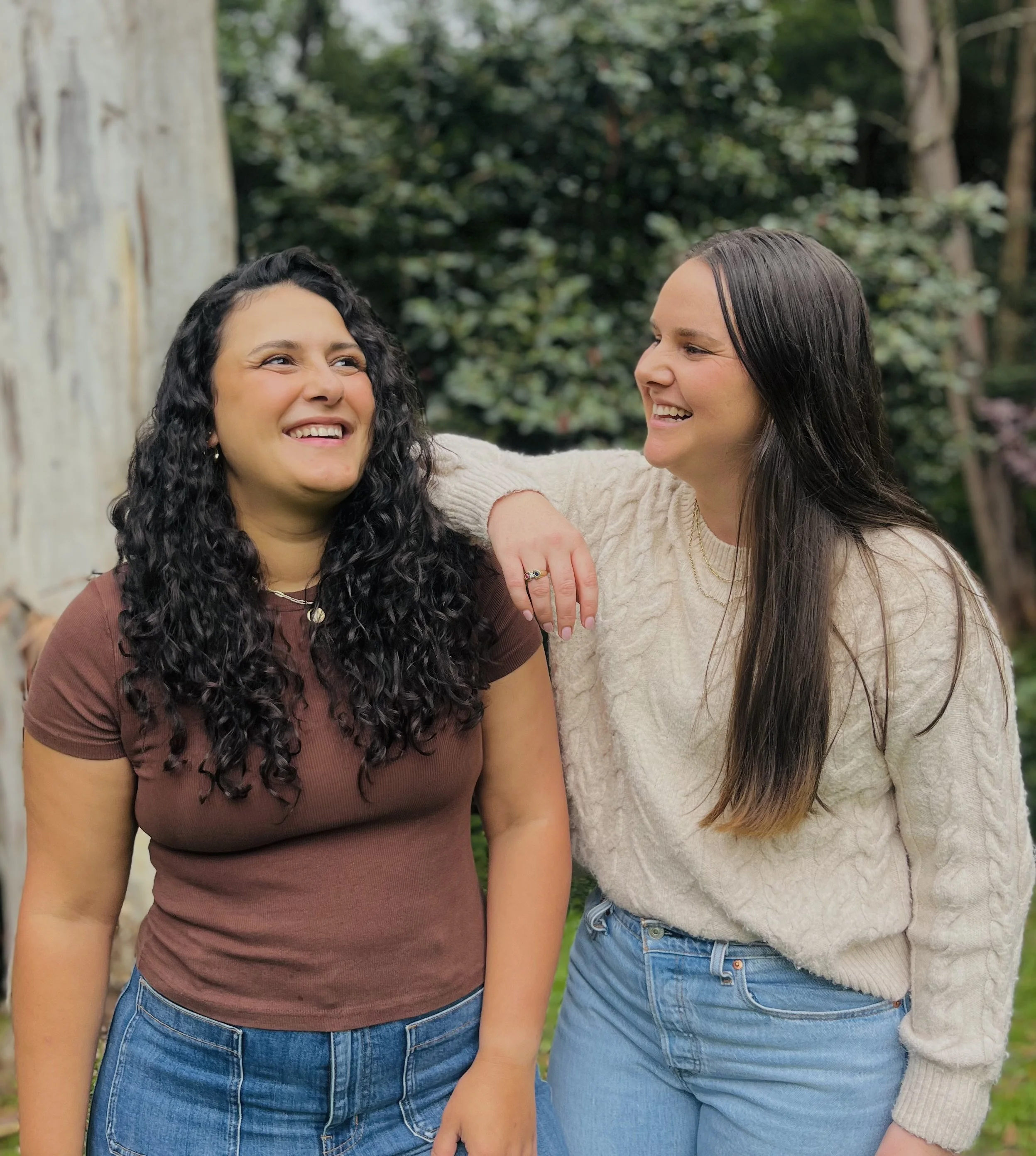 Two women smiling outdoors, one with curly black hair wearing a brown shirt, the other with straight brown hair wearing a beige sweater.