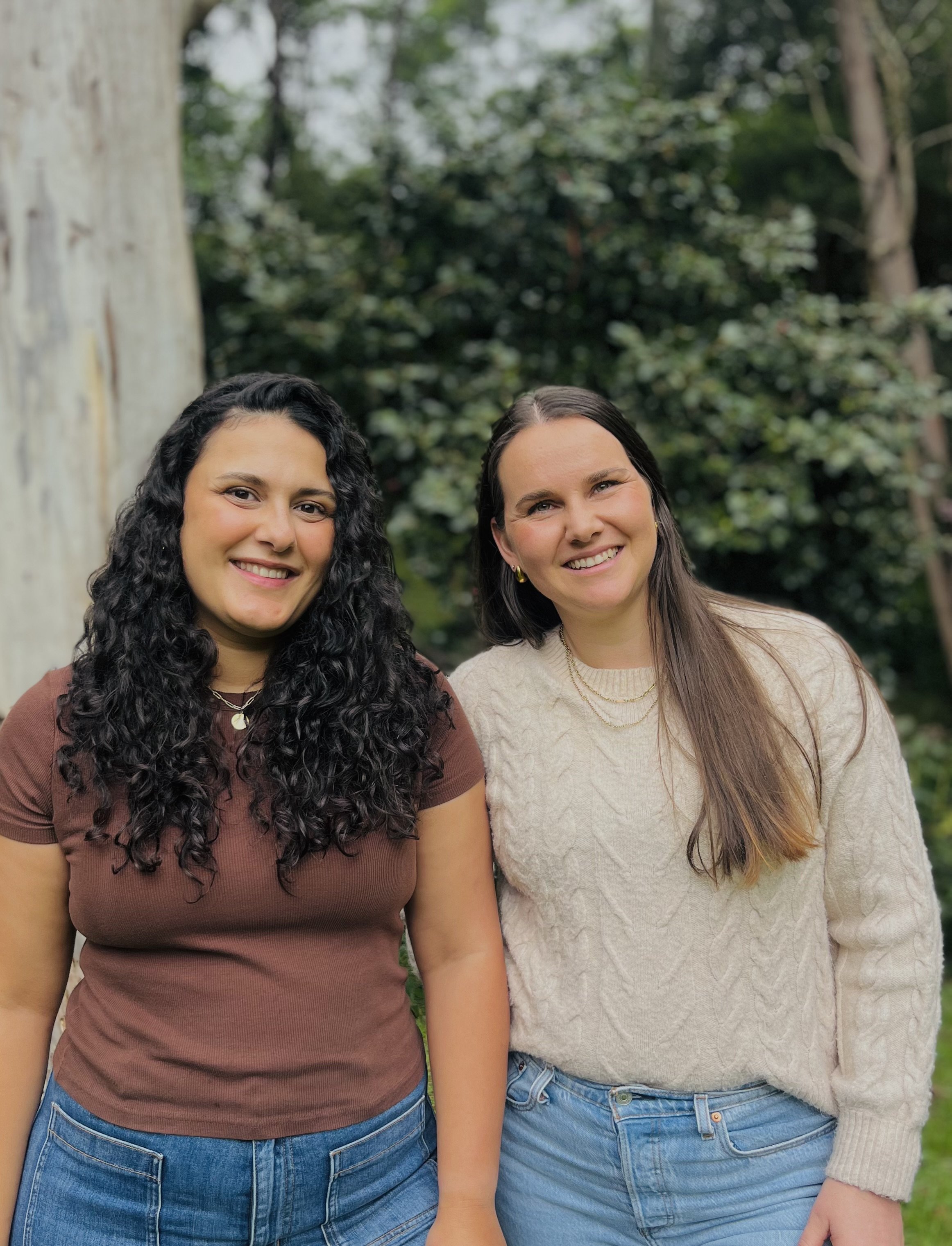 Two women standing together outdoors with a blurred leafy background, smiling at the camera.
