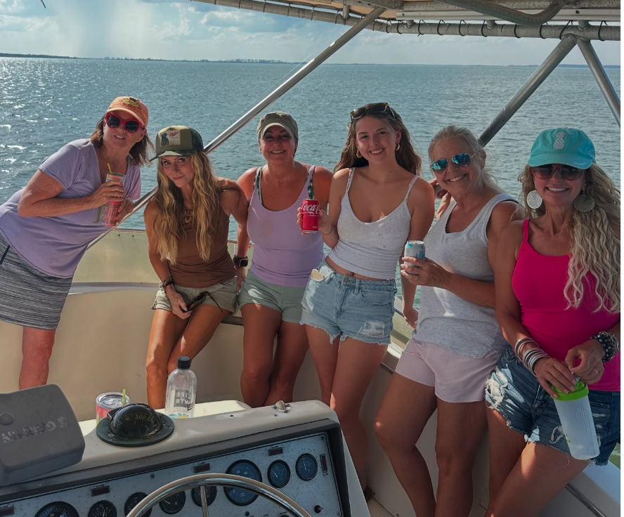 Group of six women on a boat, smiling and holding drinks, with water and a distant shoreline in the background.