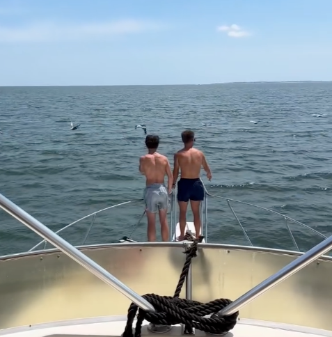 Two shirtless men in swim trunks standing on the bow of a boat, fishing in the ocean with seagulls flying above.