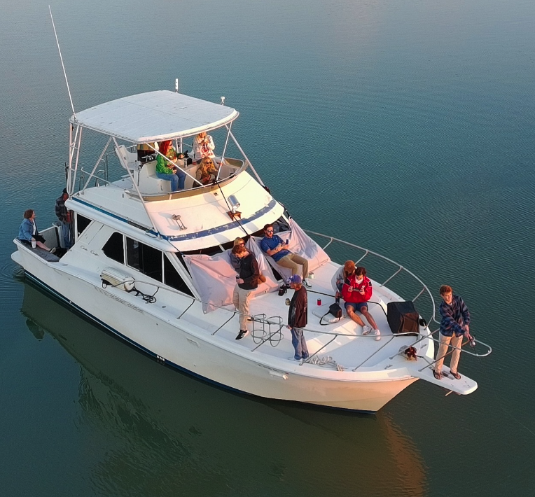 A group of people relaxing and socializing on a white yacht on calm water, with some sitting on the deck and others on the upper level, enjoying sunny weather.