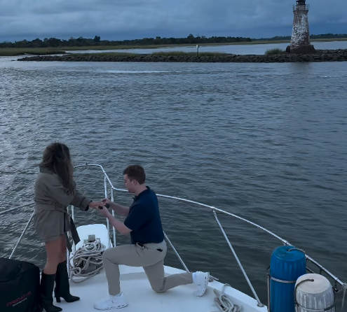 A man kneeling on a boat proposes to a woman by holding her hands, with a lighthouse and water in the background.