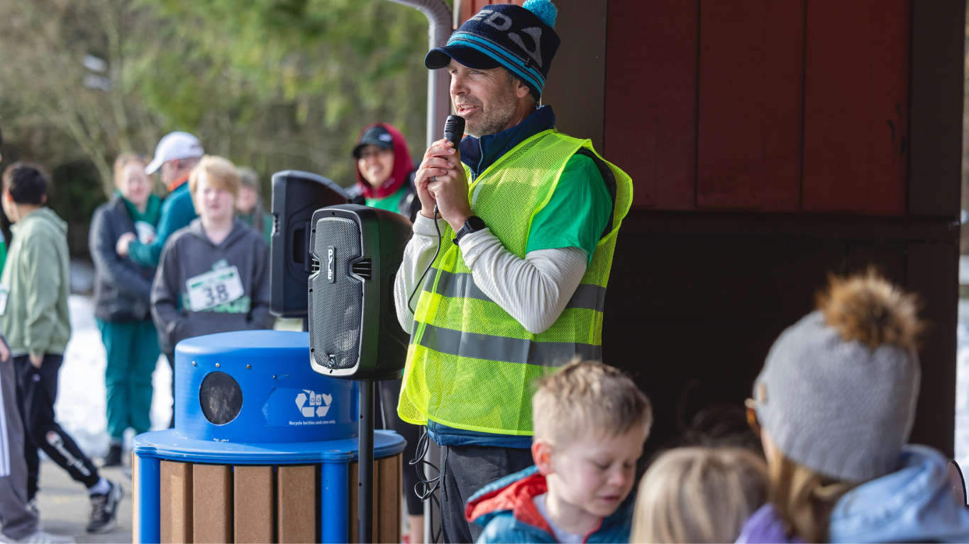 Man in yellow safety vest speaking into a microphone at an outdoor event surrounded by children and adults, with some in winter clothing and a wooded background.