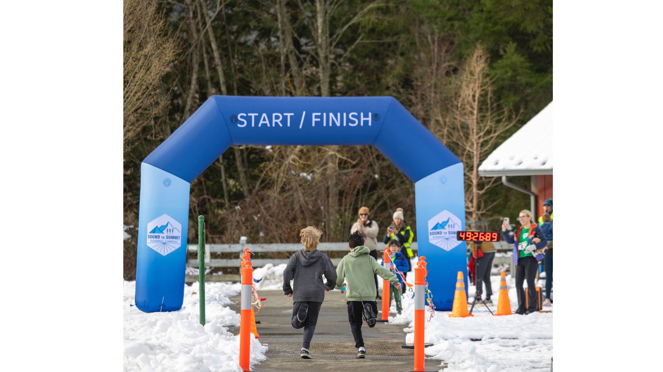 Two children running toward the start/finish line at a snowy outdoor race event, with onlookers and event staff nearby, and a digital timer showing 49:26:89.