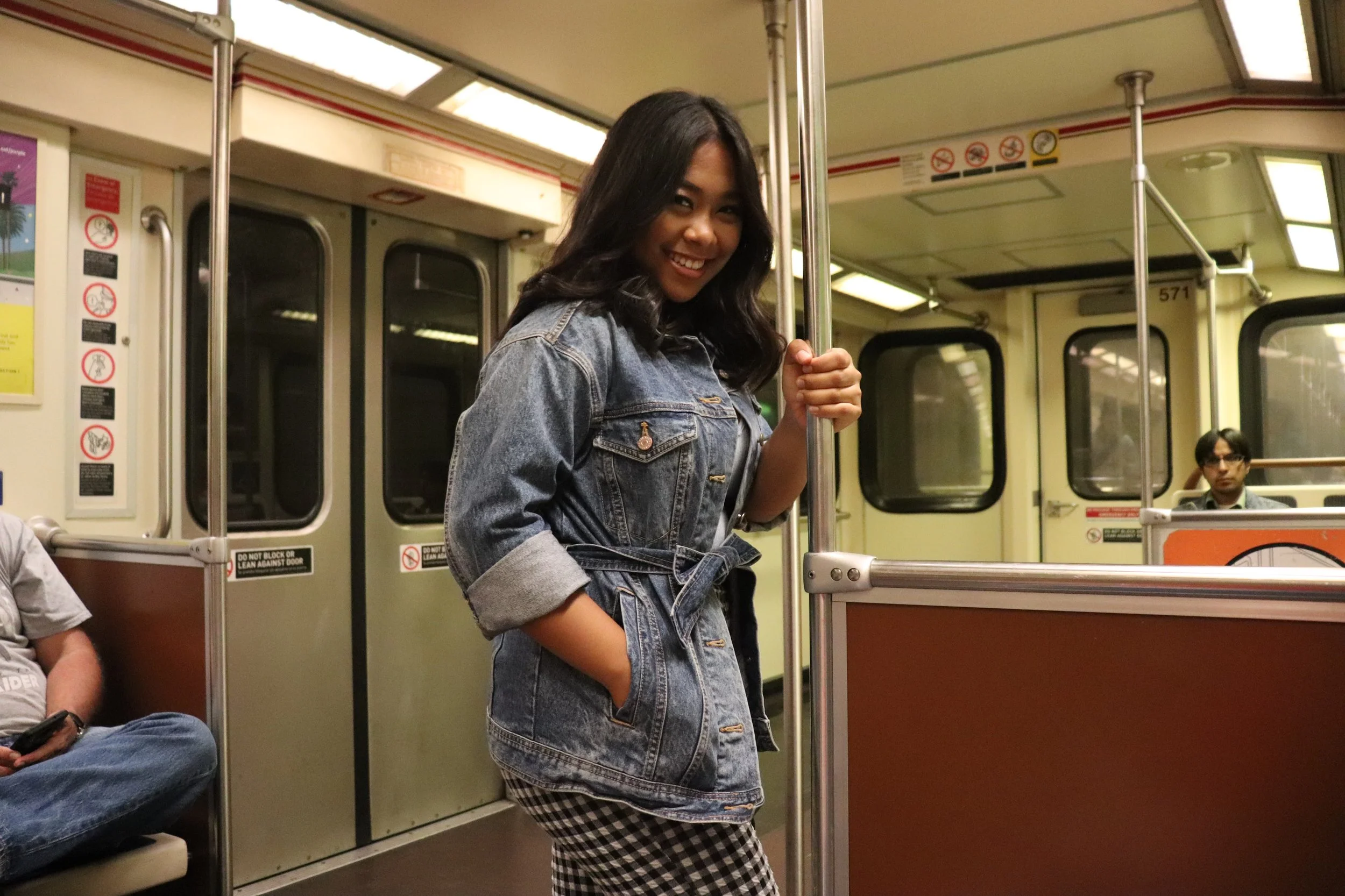A young woman with dark hair smiling on a subway train, holding a vertical pole, wearing a denim jacket and checkered pants.