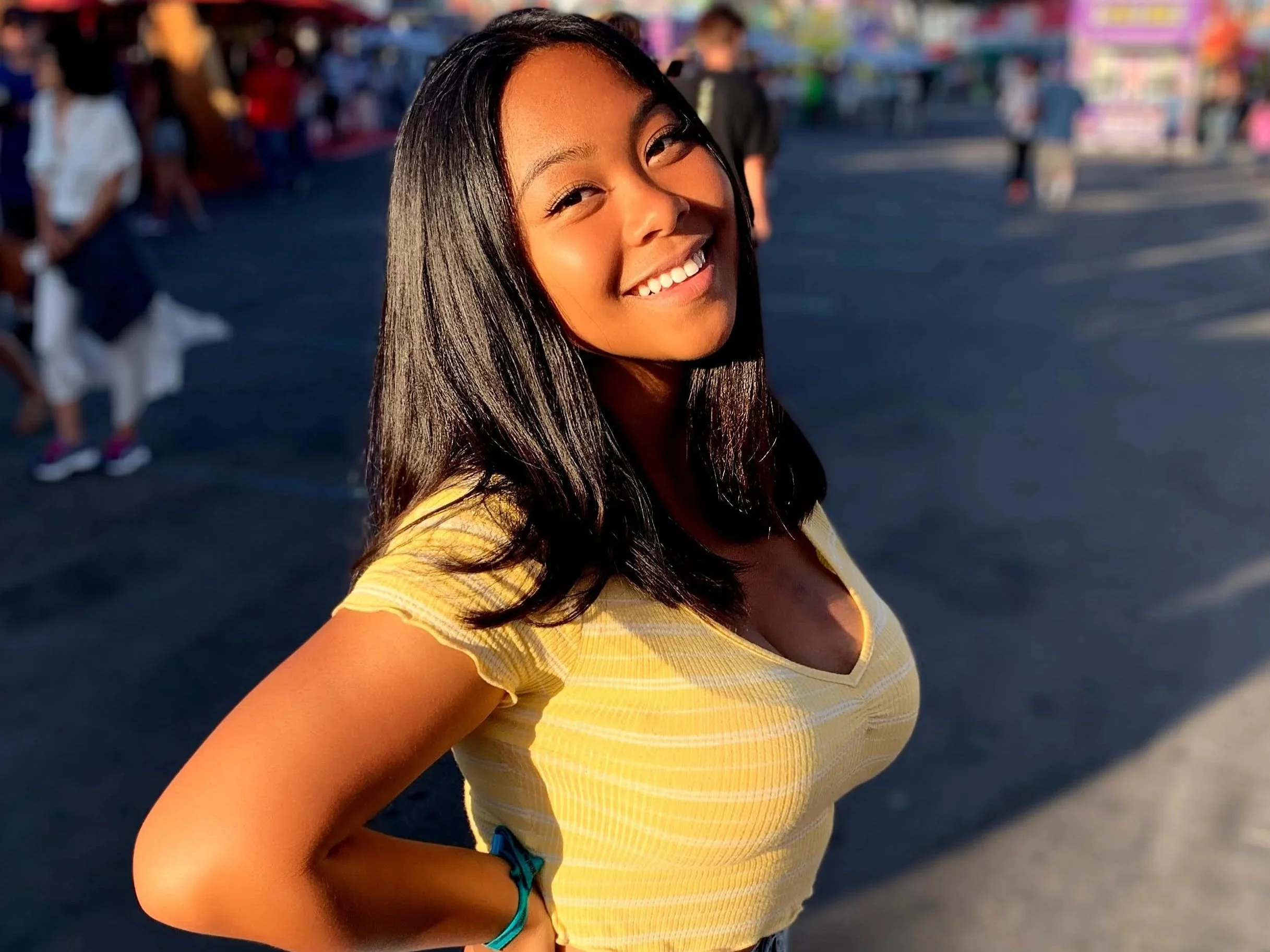 Young woman smiling, standing outdoors in sunlight, wearing a yellow top, with dark hair, at a fair or outdoor event.