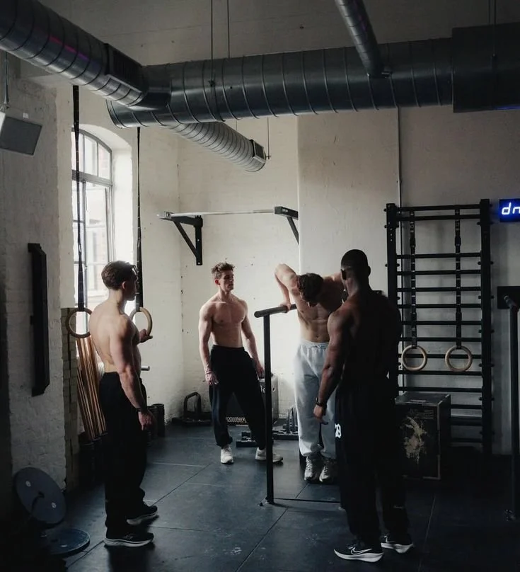 Four shirtless men with gym clothing in a fitness gym, with one man adjusting a barbell on his neck, and others standing nearby.