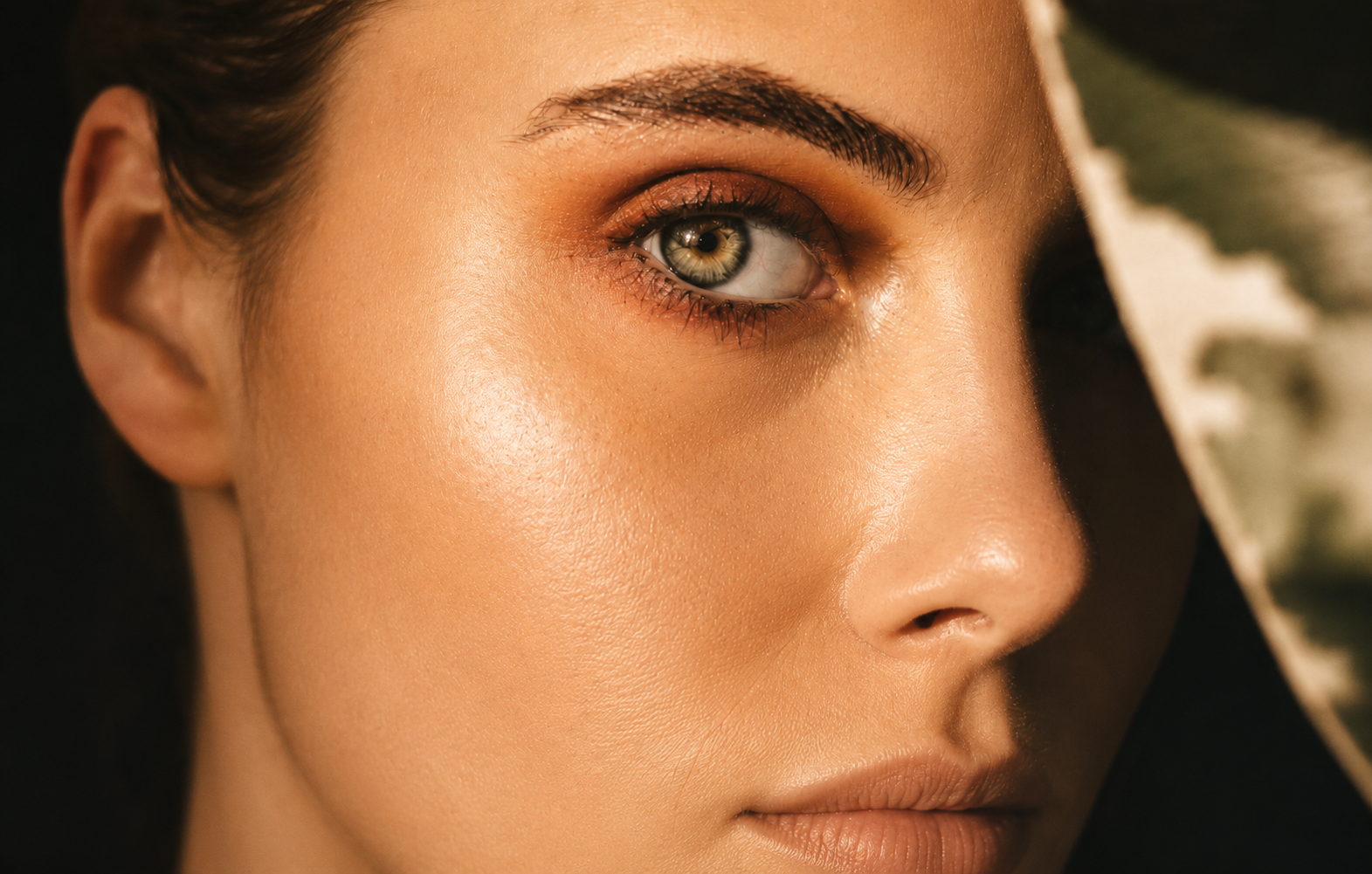 Close-up of a woman's face, focusing on her right eye with greenish-gray iris, eyebrow, and part of her nose and lips, with light makeup and smooth skin, partially shaded by a wide-brimmed hat.