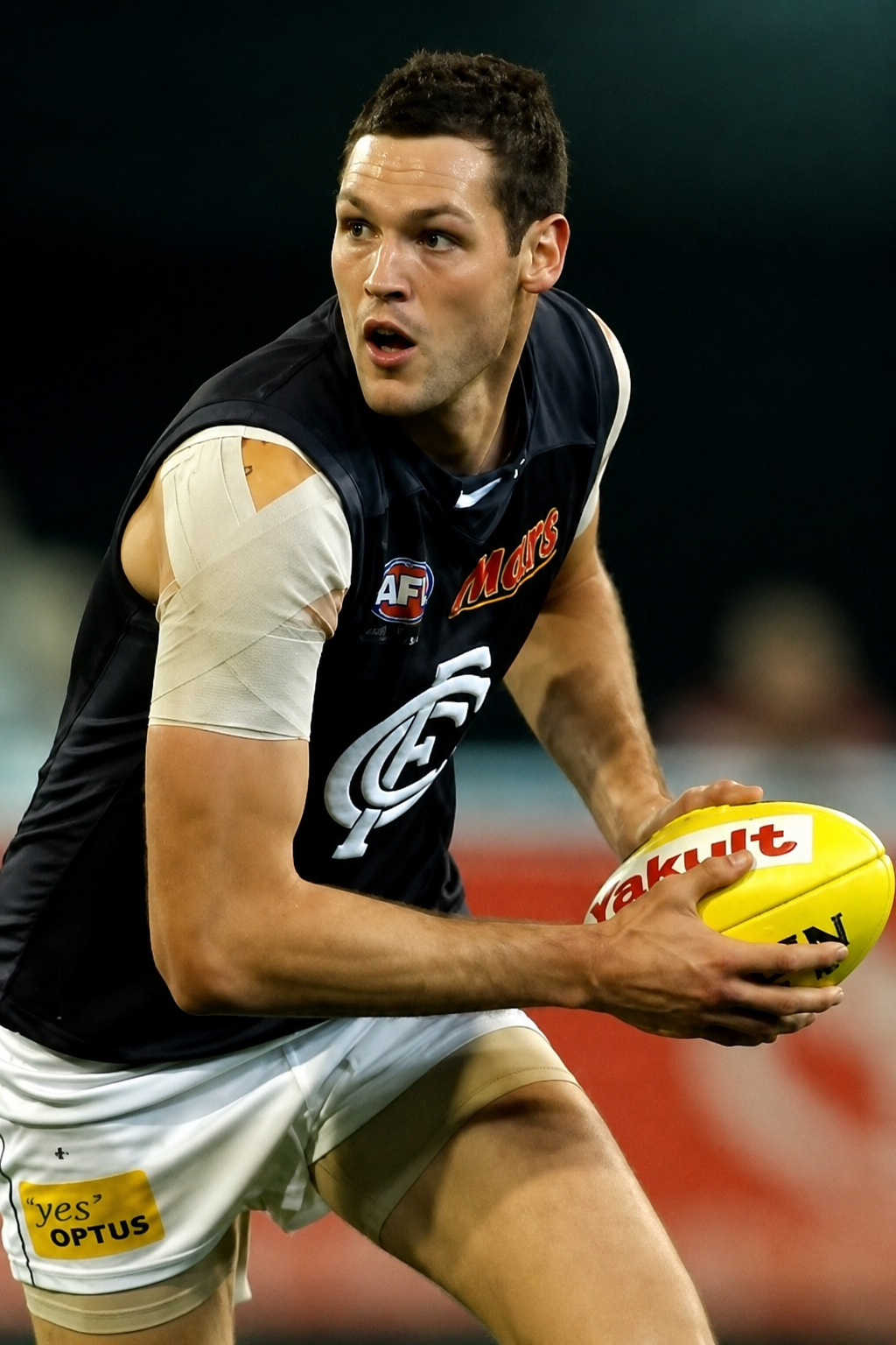 Australian football player with short dark hair, wearing a sleeveless black jersey with sponsor logos, holding a yellow football, with bandages on his upper left arm and appearing focused during a game.
