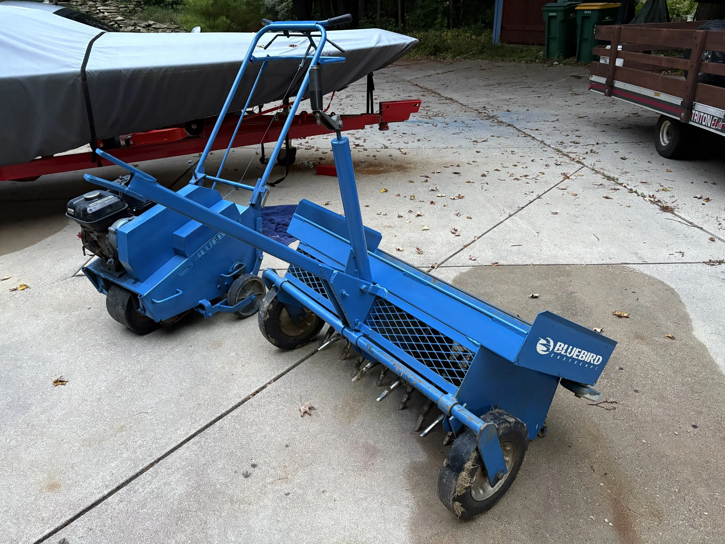 Bluebird aeration equipment on a concrete driveway next to a boat on a trailer and a utility trailer.
