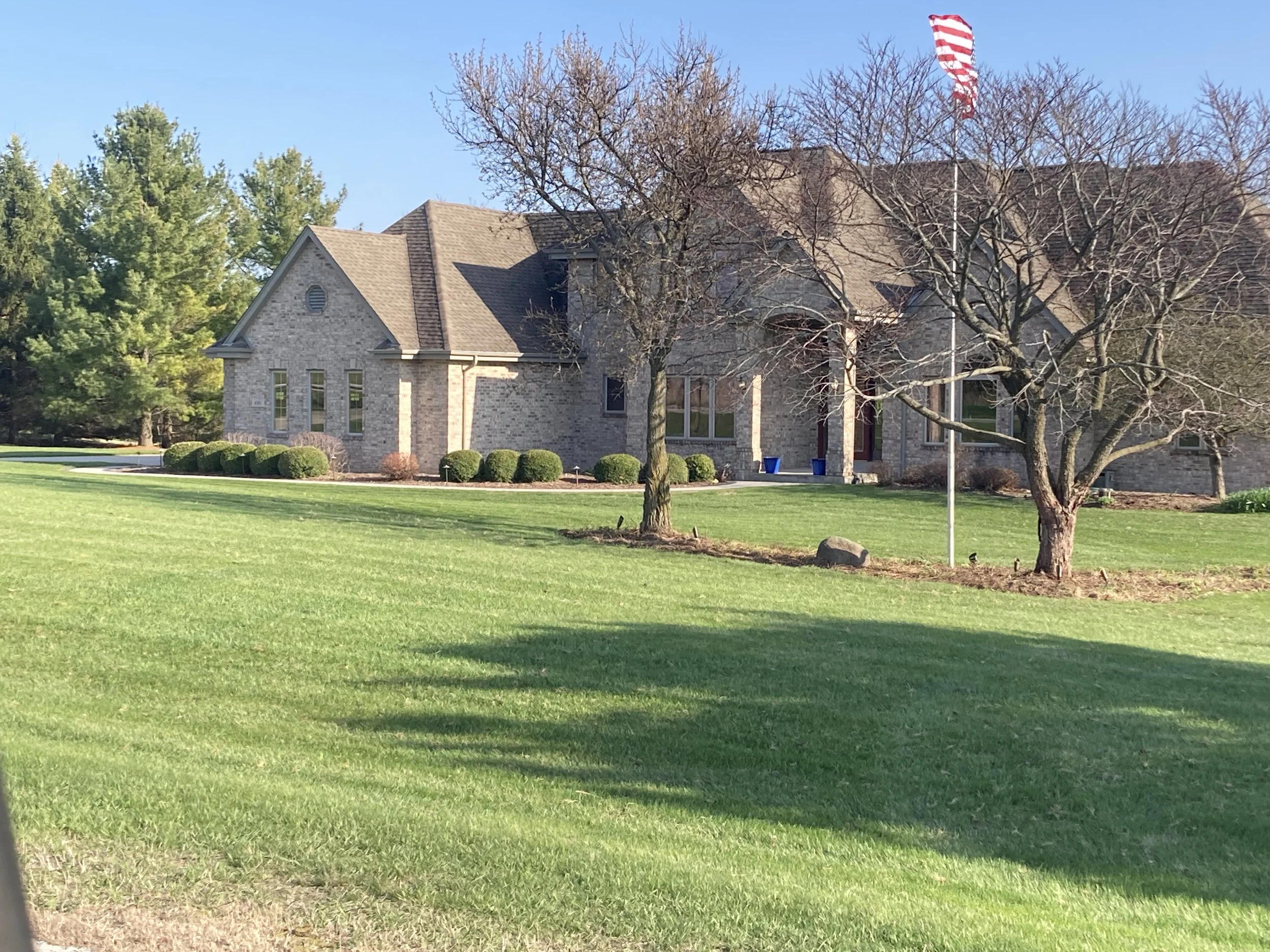 A large brick house with a steep roof, surrounded by a well-maintained lawn, leafless trees, and shrubs. An American flag is mounted on a pole in front of the house.