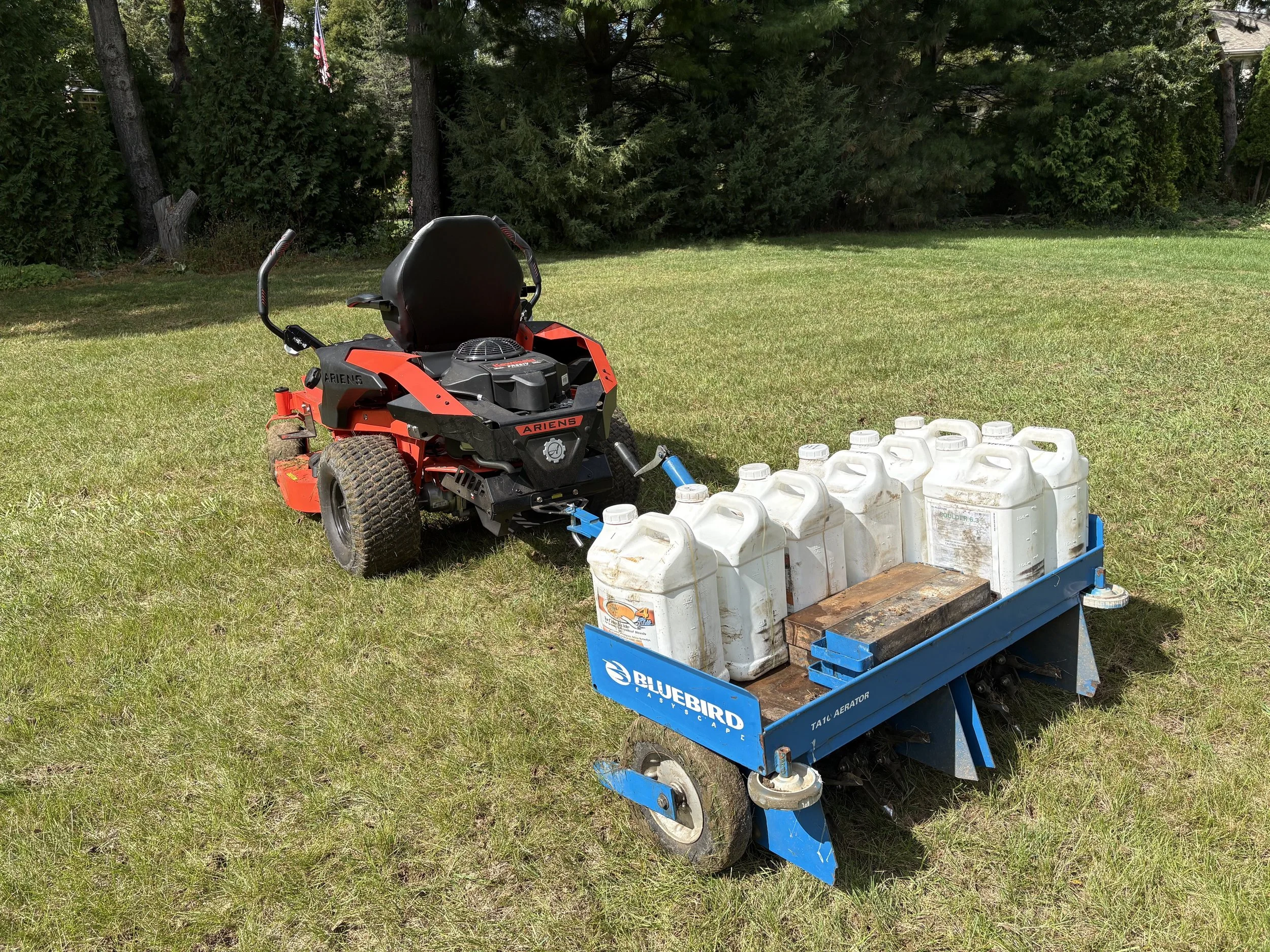 A red and black Ariens lawnmower attached to a blue Bluebird cart carrying several white plastic containers filled with liquid on a grassy lawn with trees in the background.
