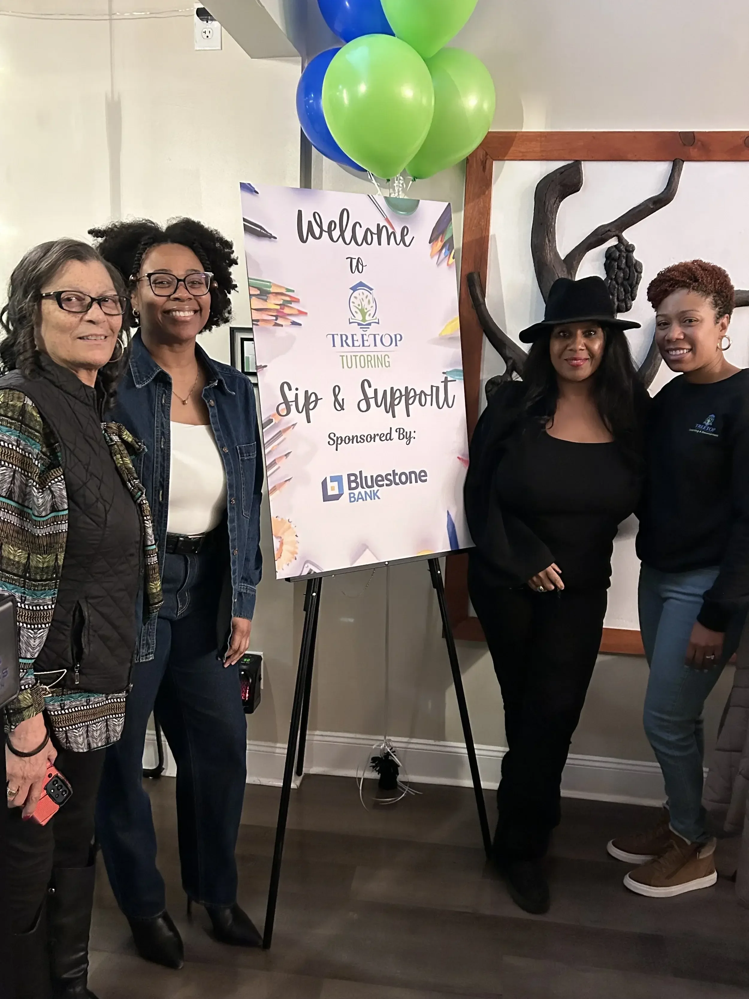 A group of four women standing near a sign that reads 'Welcome to Treetop Tutoring Sip & Support Sponsored By Bluestone Bank,' with green and blue balloons attached above the sign in an indoor setting.