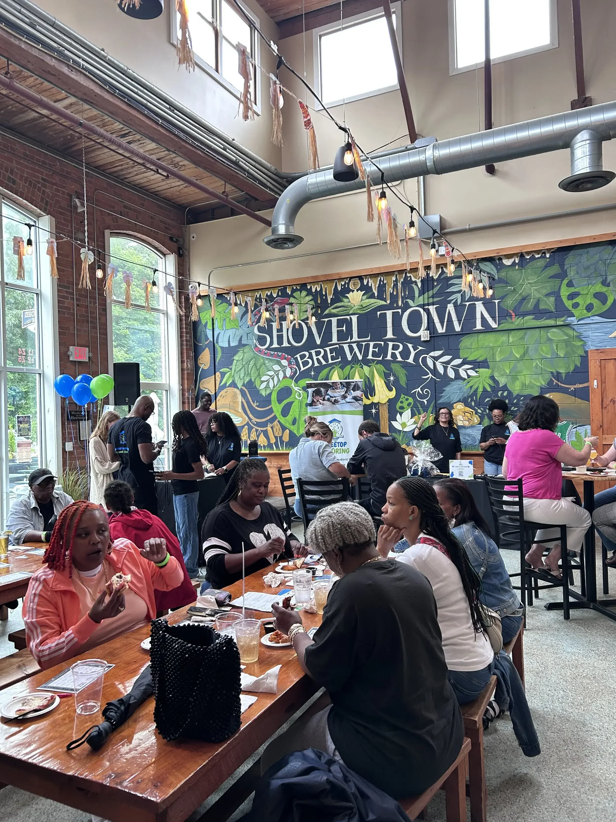 People gathered in a brewery, some sitting at long wooden tables eating pizza, others standing and chatting. The interior features high ceilings with wooden beams, brick walls, and large windows. There is a colorful mural on one wall that reads 'Shovel Town Brewery' with greenery and whimsical designs. Blue and green balloons are visible, and string lights with tassels hang from the ceiling.